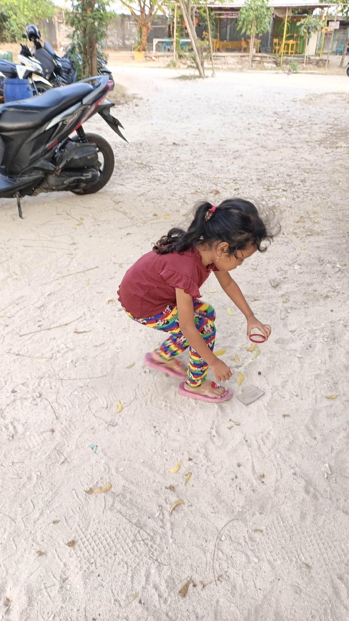 A young girl with dark hair in a ponytail, wearing a red shirt and brightly colored pants with a Mickey Mouse print, is crouched on sandy ground. She is wearing pink flip-flops. In her right hand, she holds a small, red, circular object. To her right, on the ground, is a small, grey, rectangular object. The ground is covered in sand, with scattered leaves and some tire tracks visible. In the background, a black scooter is parked, and beyond that, a building with an open-air structure, possibly a market or restaurant, is visible with trees and other structures. The overall setting appears to be an outdoor area in a tropical or subtropical location, possibly a village or a less developed part of a city, consistent with Cikal, Indonesia. The lighting suggests it is daytime, with the sun casting some shadows.