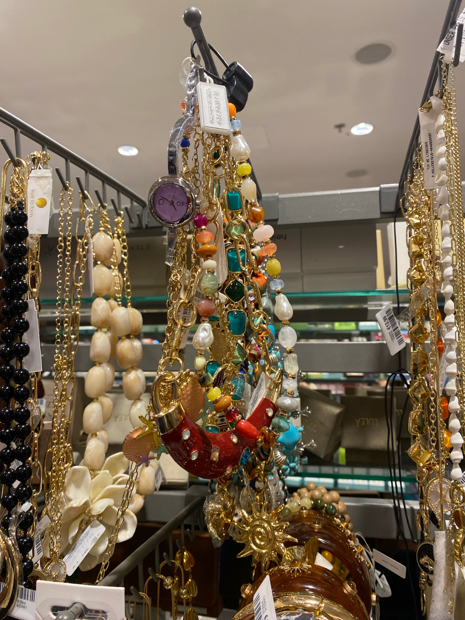 The image shows a close-up, eye-level, indoor shot of a display rack filled with various necklaces and bracelets. The items are primarily gold-toned and feature an assortment of colorful beads, charms, and pendants, including stones, pearls, and geometric shapes. Among the jewelry, a small purple-faced watch with a silver band is visible. Several white price tags with barcodes and text, some in German, are attached to the jewelry. The background consists of shelves with boxes and more retail merchandise, suggesting a store environment. The lighting is bright, likely from overhead store fixtures, creating some glare on the metallic elements of the jewelry. The specific location in Vienna, Austria cannot be determined from the image alone, as there are no identifiable landmarks or signage that indicate the city.