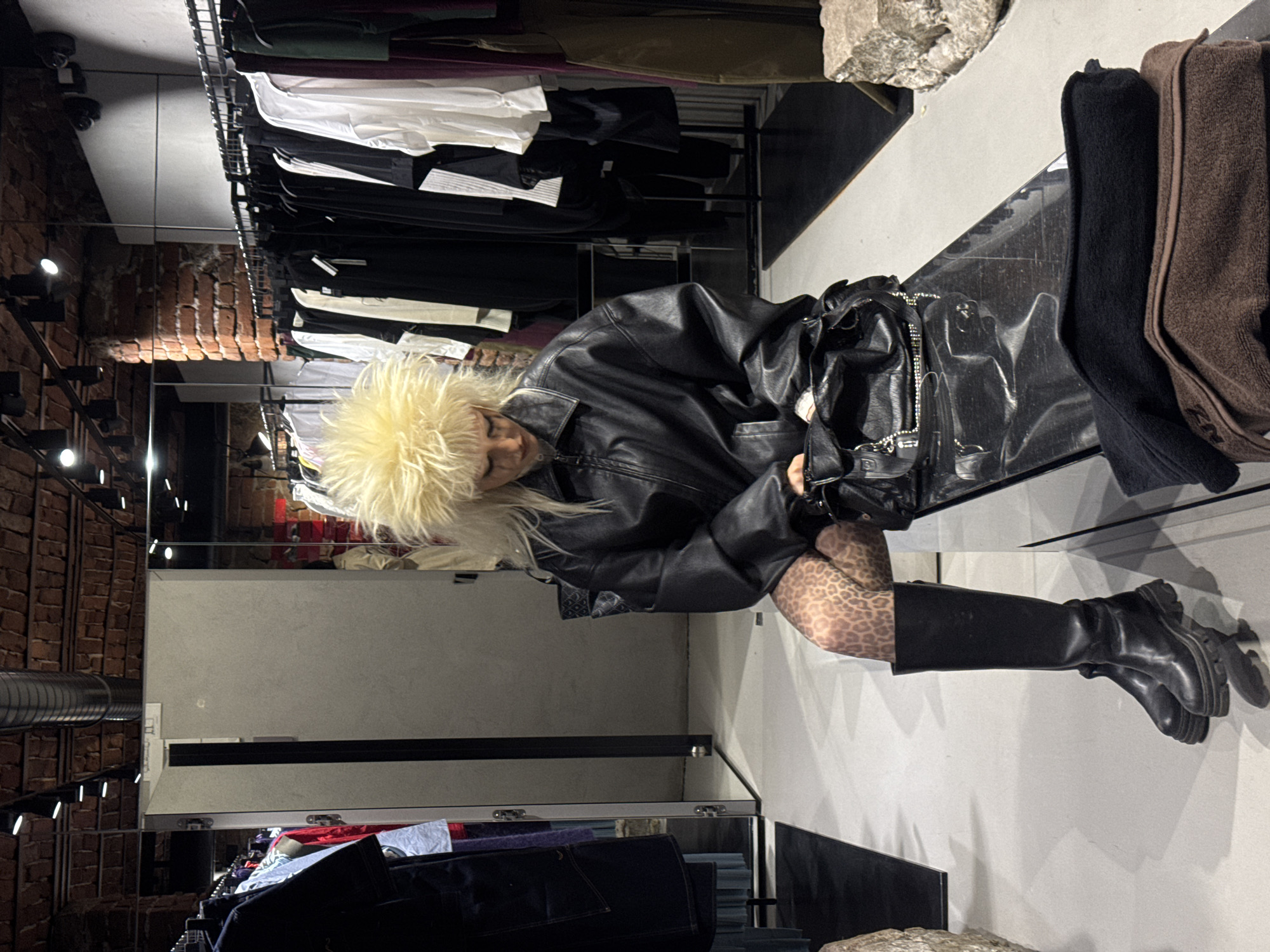 The image depicts a modern clothing boutique with an industrial aesthetic, identified as Collective Store, likely located on Tsar Shishman Street in Sofia, Bulgaria.
A single individual is centrally positioned, seated on a dark, reflective bench. This person has voluminous, spiky blonde hair and wears an oversized black leather bomber jacket, leopard print tights, and tall black lug-soled boots. Their head is tilted downwards, and they are looking into a black, textured handbag placed on the bench, with their hands near its opening. Distinct eyeliner marks are visible below the eyes.
The background features exposed red brick walls and ceiling, complemented by visible silver ductwork and black track lighting fixtures. Multiple clothing racks, laden with garments primarily in black, white, and dark hues (including deep purple and olive), line the walls. A light-grey partition, possibly a changing room door, is visible on the left, adjacent to a large mirror. The floor is light grey and reflective. A stack of folded black and brown textiles rests on the bench to the right of the handbag, and a rough-hewn, light-colored stone block is present in the foreground on the right.