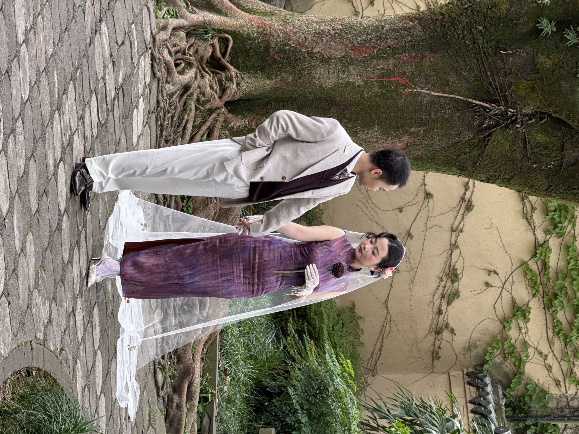A man and woman are standing outdoors on a stone-paved walkway, holding hands. The woman is wearing a purple dress and a white veil. The man is wearing a light-colored suit, vest, and white pants. They are standing near a large tree with visible roots, and there is a wall with vines growing on it in the background. The scene appears to be in a garden or park-like setting. The location is within the city of Yuzhong District, China.