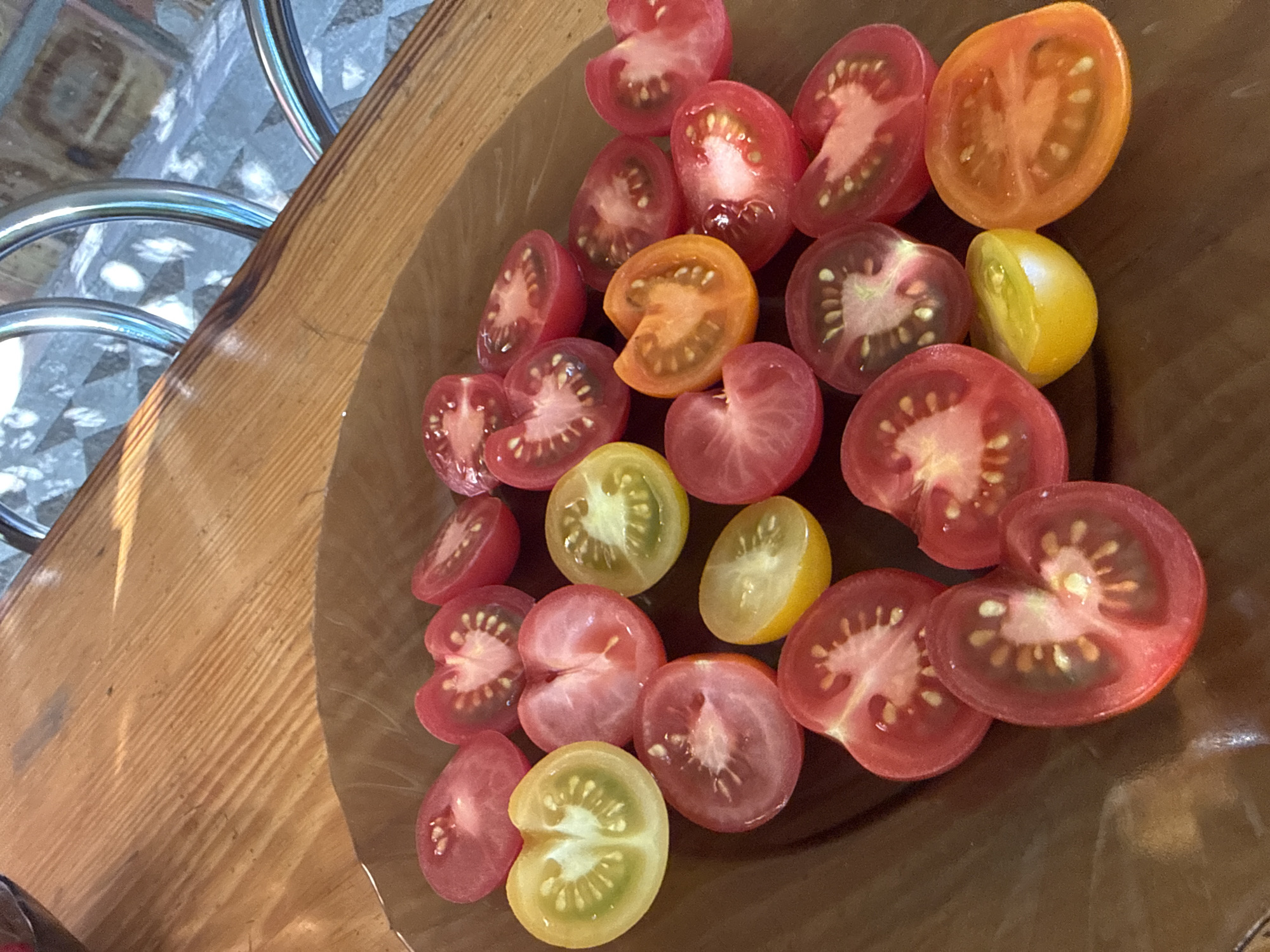 A clear, overhead shot captures a collection of halved cherry tomatoes in a clear glass bowl, resting on a wooden surface. The tomatoes display a variety of colors, including vibrant reds, oranges, and yellows. Some are sliced horizontally, revealing their juicy interior and seeds, while others are cut vertically, showcasing their delicate shapes. The wooden surface appears to be a table or countertop with visible grain patterns and a light, natural finish. In the background, a blurred metallic structure, possibly part of a chair or table, adds a subtle contrast to the organic shapes of the tomatoes. The lighting suggests it could be daytime, with natural light casting soft shadows and highlights on the tomatoes and the bowl. The overall impression is one of freshness and preparation for a meal.