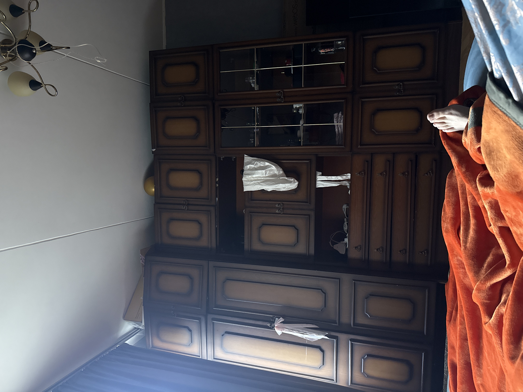 The image depicts a dimly lit bedroom interior. In the foreground, an orange blanket is draped over a person's legs and feet, with one bare foot visible. Behind the bed, a large, dark wood wardrobe with multiple doors and drawers dominates the scene. Some doors have glass panes revealing items stored inside, while others are solid. A white fabric hangs from one of the solid doors. Above the wardrobe, a portion of the ceiling with light fixtures is visible. The overall atmosphere is domestic and casual. The location is specified as Moscow, Russia.