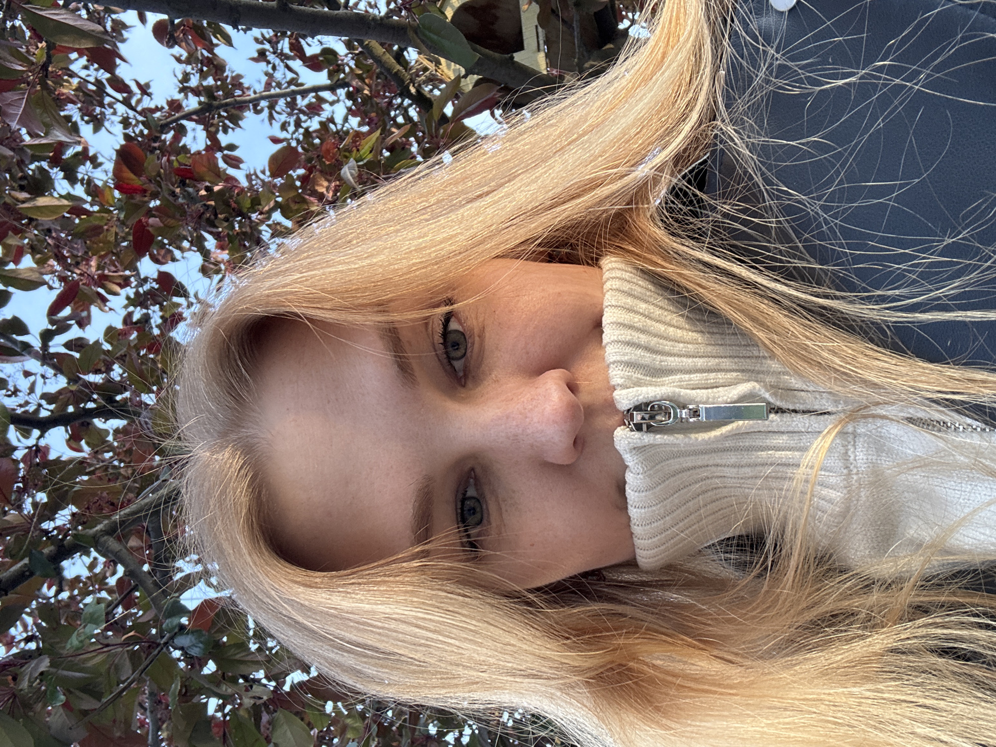A young woman with long blonde hair is facing the camera. She is wearing a light-colored, zipped-up sweater under a dark-colored jacket. The background shows tree branches with red and green leaves against a blue sky.
The exact location in the city of Podosenikha, Russia, cannot be determined from the image.