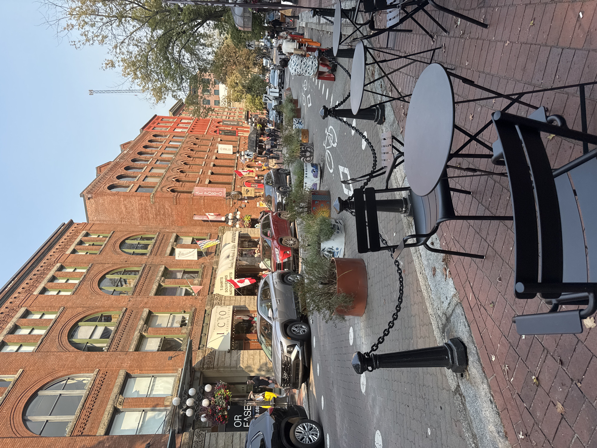 This image depicts a street scene in Gastown, Vancouver, Canada. The street is paved with bricks and lined with heritage buildings made of red brick. The buildings have arched windows with green trim. Several people are visible walking on the sidewalk and in the street. Cars are parked along the street and driving. There are outdoor seating areas with tables and chairs set up on the sidewalk. Planters with greenery are placed along the street. The sky is clear and blue. There is a cellular tower visible in the distance.