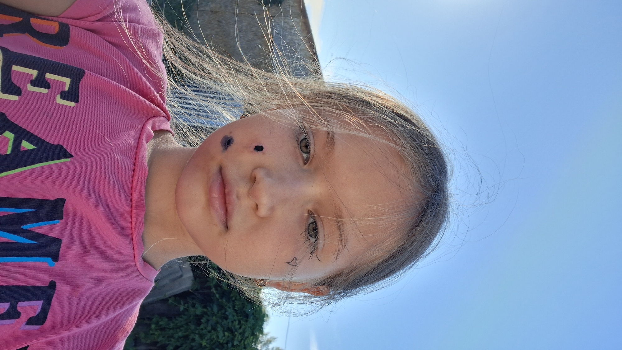 A young girl with blonde hair and light skin is shown from the chest up, looking directly at the camera. She has several dark markings on her left cheek and a small heart drawn on her right cheek. She is wearing a pink t-shirt with colorful lettering. The background appears to be outdoors with a bright blue sky and some greenery. The overall lighting suggests it is daytime. There is no discernible location in the city of Semey, Kazakhstan.