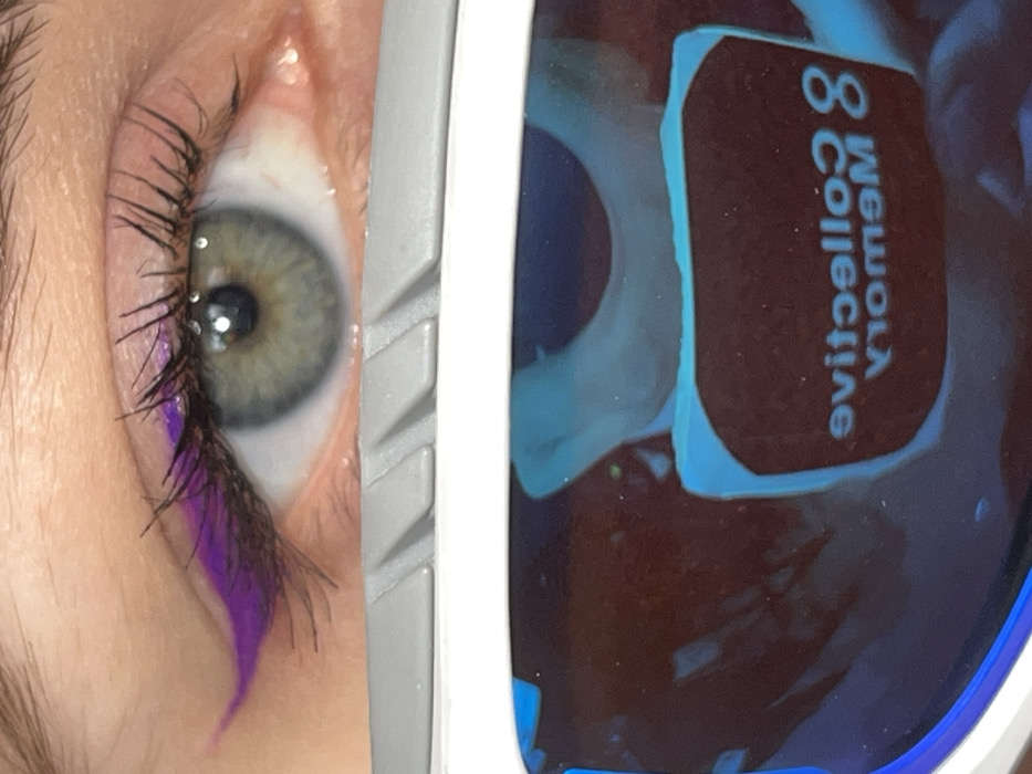The image presents a close-up, downward-angled view of a human right eye with elaborate makeup, positioned above a reflective, framed surface. The eye features an iris of green and hazel tones with a dark pupil. The upper eyelid is adorned with a prominent, sharp winged eyeliner in a vibrant purple hue, extending towards the outer corner. Dark mascara is applied to the eyelashes. A portion of a light brown eyebrow is visible at the top edge of the frame. Directly below the eye is a grey, rigid object with parallel ridged details, likely the frame of glasses or a digital viewing device. Below this frame, the lower half of the image displays a dark, highly reflective surface, possibly a screen or lens. This surface reflects luminous blue, abstract shapes, and mirrored white text which reads "8 collective Memory", indicating the actual text is "Memory Collective 8". The scene is static and appears to be an indoor setting, possibly in Krasnoyarsk, Russia.