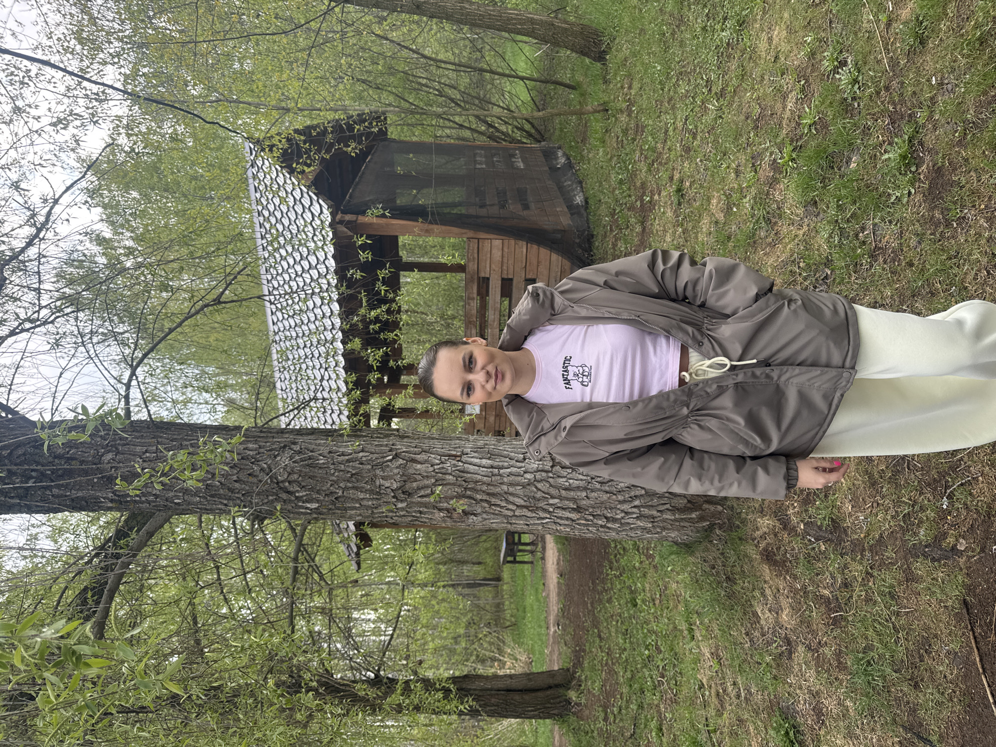 A young woman is standing in a wooded area, in Chaikovsky, Russia. The woman is wearing a gray-green jacket over a light pink t-shirt and white pants. She is positioned next to a large tree. Behind her is a wooden structure with a mesh screen roof. The ground is covered in grass and fallen leaves. Trees surround the scene.