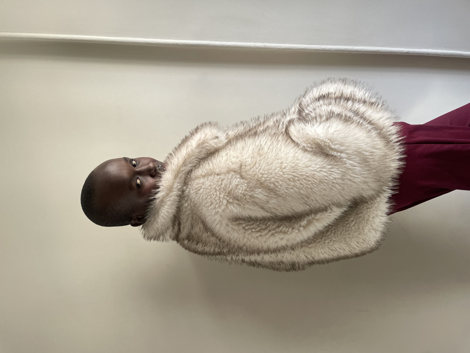 The scene is set in an indoor location in Paris, France. A dark-skinned individual with a shaved head is depicted from the upper torso, turned away from the camera, looking back over their left shoulder. They are wearing a fluffy, multi-toned faux fur coat, predominantly white or cream with irregular brown or grey horizontal stripes, which covers their upper body. A portion of deep red fabric, consistent with pants or a skirt, is visible below the coat on the right side. The individual's face is directed towards the viewer, exhibiting a subtle expression and a small metallic nose ring on their right nostril. The background is a plain, light-colored wall, featuring a vertical architectural trim or panel on the right side of the frame. The lighting is soft and ambient.