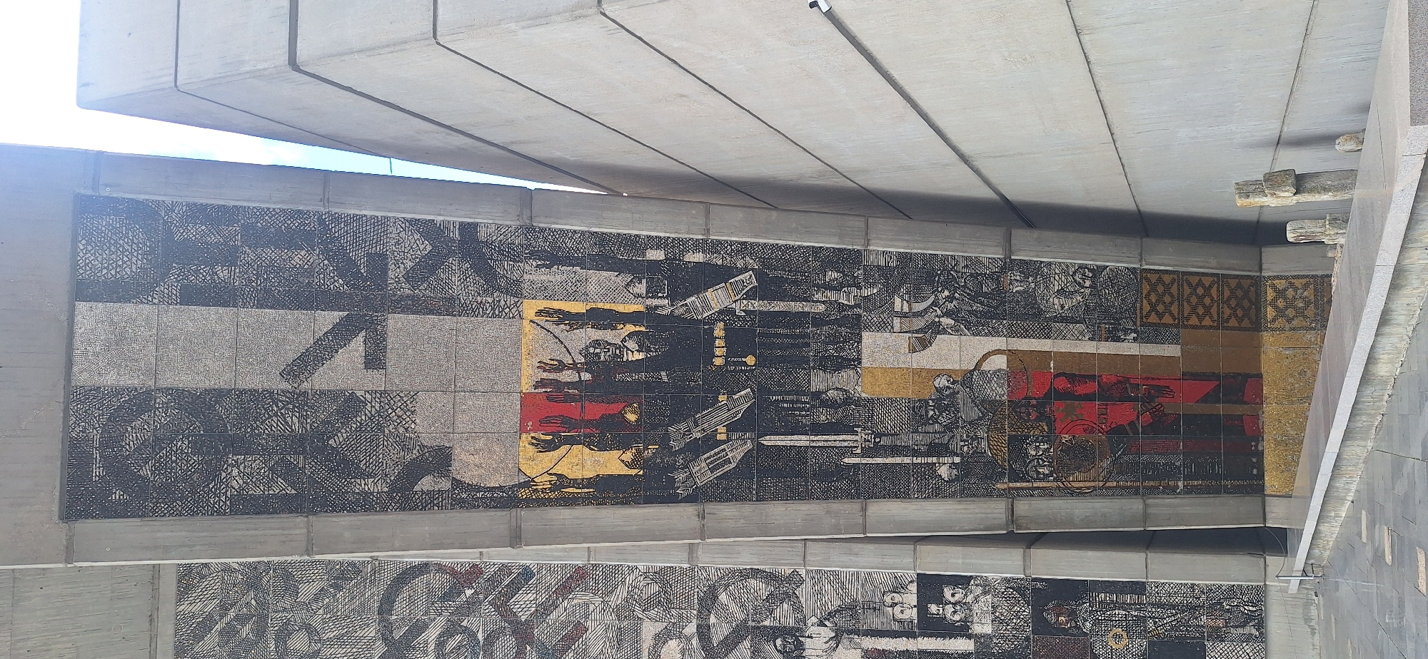 The image shows a large mosaic mural on a concrete structure in Shumen, Bulgaria. The mosaic is composed of many small tiles and depicts a scene with several figures. In the center of the mural, a group of people with raised arms are shown against a backdrop of red and yellow tiles. They are wearing dark clothing and some appear to be holding weapons or tools. The surrounding areas of the mosaic feature abstract patterns and other figures that are less distinct. To the right of the mosaic, a plain concrete wall rises at an angle, and to the left, another section of a mosaic mural is visible. The ground in front of the structure is paved with gray tiles. A low concrete ledge runs along the front of the mural, and a few small stone objects are placed on it. The lighting suggests it is daytime, with a bright sky visible in the upper right portion of the image.
