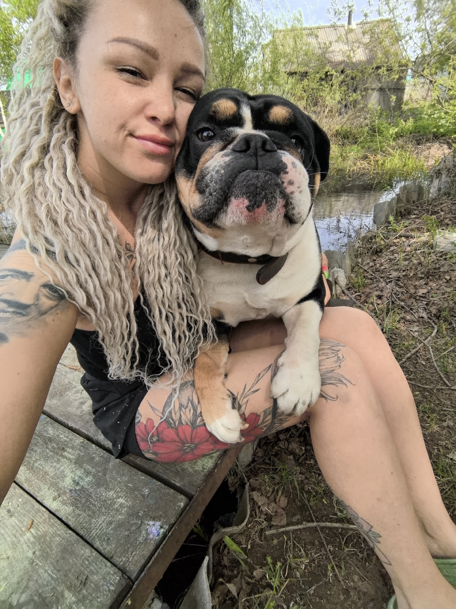 A woman with long, light-colored dreadlocks is holding a bulldog on her lap. She is sitting on a weathered wooden platform, with a body of water and greenery visible in the background. The woman has tattoos on her arms and legs. The dog wears a dark collar. The scene appears to be outdoors, possibly near a river or pond. It is not possible to determine if the location is in the city of Noviy, Russia based solely on the image.