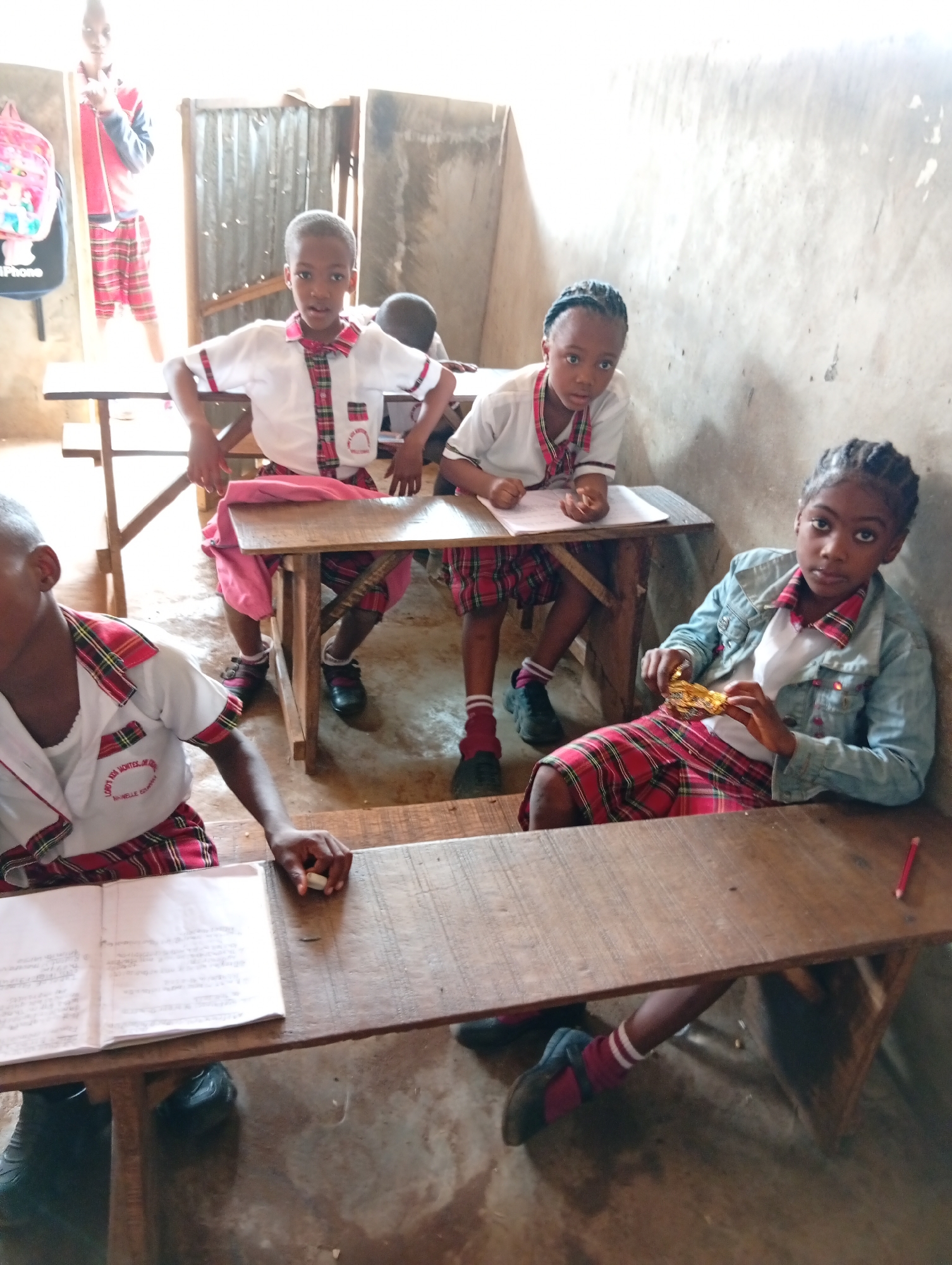 In what appears to be a classroom setting in Ukwu-Abwa, Nigeria, several children are seated at wooden desks. The children are wearing school uniforms consisting of white shirts, red and black plaid bottoms, and black shoes. They are situated in a room with concrete walls and a metal door. The children are interacting with school books and writing implements on their desks. Another child in similar clothing is standing in the background, in the doorway with a backpack.