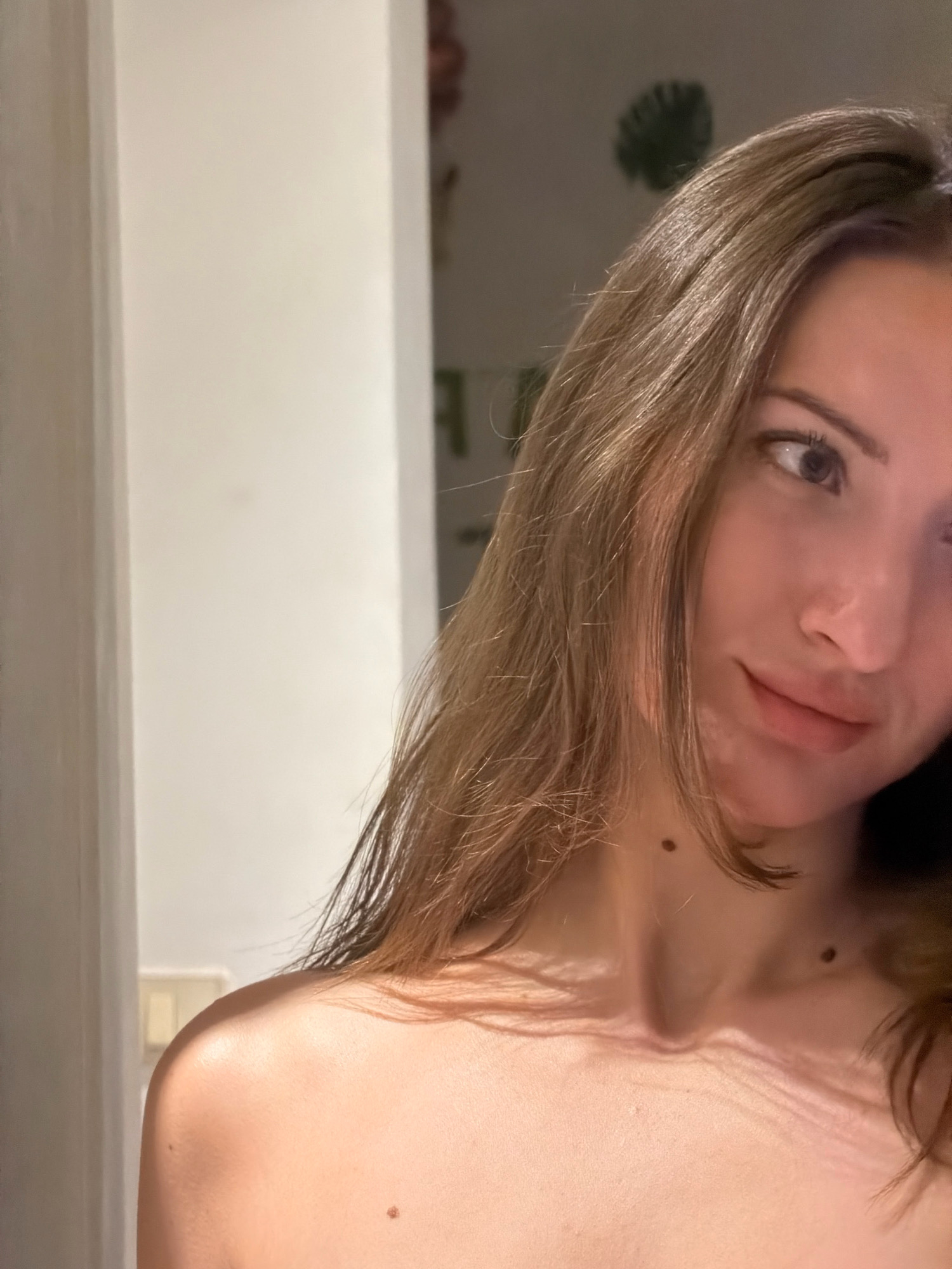The image shows a close-up, angled selfie of a young Caucasian woman. She has long, light brown hair that frames her face and falls over her shoulders. Her gaze is directed away from the camera, towards the right. She is bare-shouldered, and the lighting casts a warm glow on her skin, highlighting her collarbones and shoulders. A mole is visible on her neck. The background is softly blurred, with hints of light-colored walls and possibly some decorative elements such as a plant silhouette. The overall impression is an intimate and softly lit portrait. The image provides no information about its location in Sharm El Sheikh, Egypt.