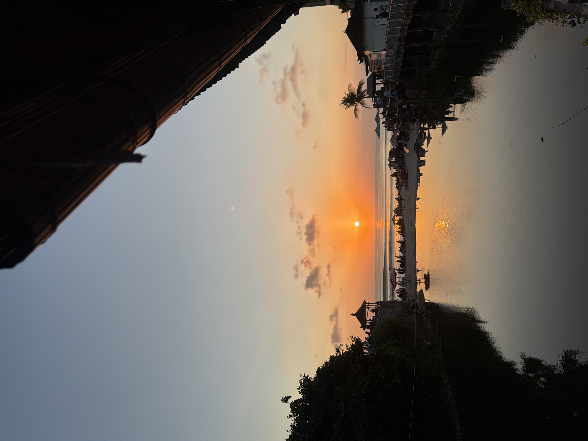 This is a vertical, eye-level shot of a sunset over a beach and a river in Kuta Selatan, Indonesia, likely at the scenic Melasti Beach. The sky is a gradient of pale blue at the top transitioning to a vibrant orange as it nears the horizon. The sun, a bright orange orb, is low on the horizon, casting a warm glow and reflecting on the water.
In the foreground, a dark body of water, possibly a river or estuary, fills the lower half of the frame. The water's surface is calm and reflects the colors of the sunset and the silhouettes of the structures and trees on the shore.
On the left bank of the water, a dense, dark green tree is visible, its leaves obscuring part of the view. Adjacent to the tree, a stone structure with a traditional Balinese-style gazebo or pavilion sits atop a rocky outcrop. Several figures are silhouetted on this outcrop, appearing to observe the sunset.
Across the water and further down the beach, a sandy stretch is dotted with people. Umbrellas and small tables suggest a beachside dining or lounging area. Several structures, appearing to be cafes or restaurants with thatched roofs, are built on stilts over the water or along the edge of the beach. A single palm tree stands tall near one of these structures. The ocean in the background shows gentle waves.
The top right of the image is dominated by a dark, diagonal structure, possibly the roof of a building or a viewing platform, made of natural materials like bamboo or wood, casting a shadow over the scene. The overall atmosphere is serene and picturesque, capturing the beauty of a tropical sunset.