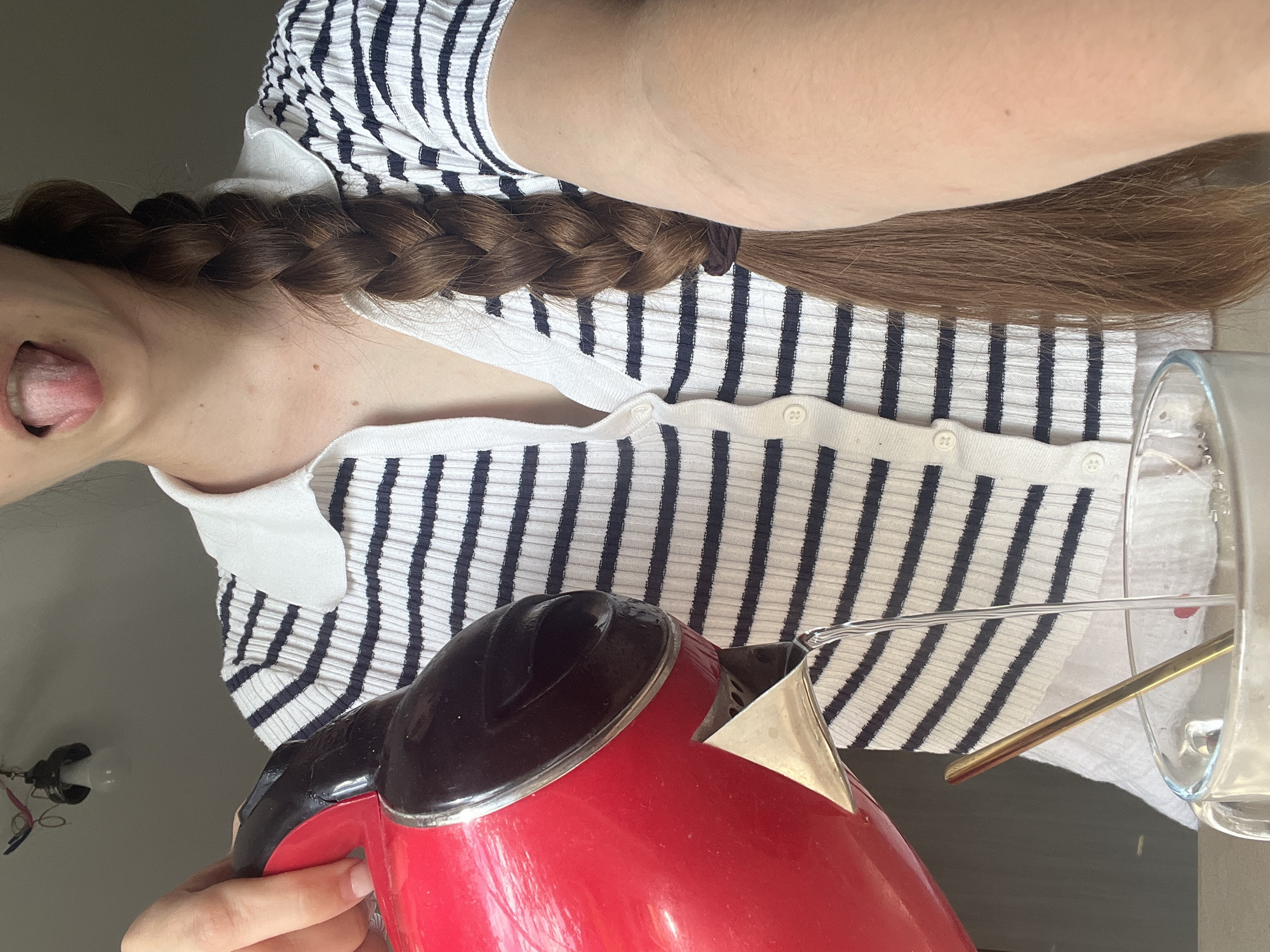 A close-up shot shows a person with a long braid of brown hair pouring hot water from a red electric kettle into a clear glass mug. The person is wearing a white collared shirt with thin navy blue stripes and has their tongue sticking out. A golden stirring spoon is visible in the mug. The background is plain and gray, with a hint of a light fixture at the top. The image is taken from a medium-low angle.