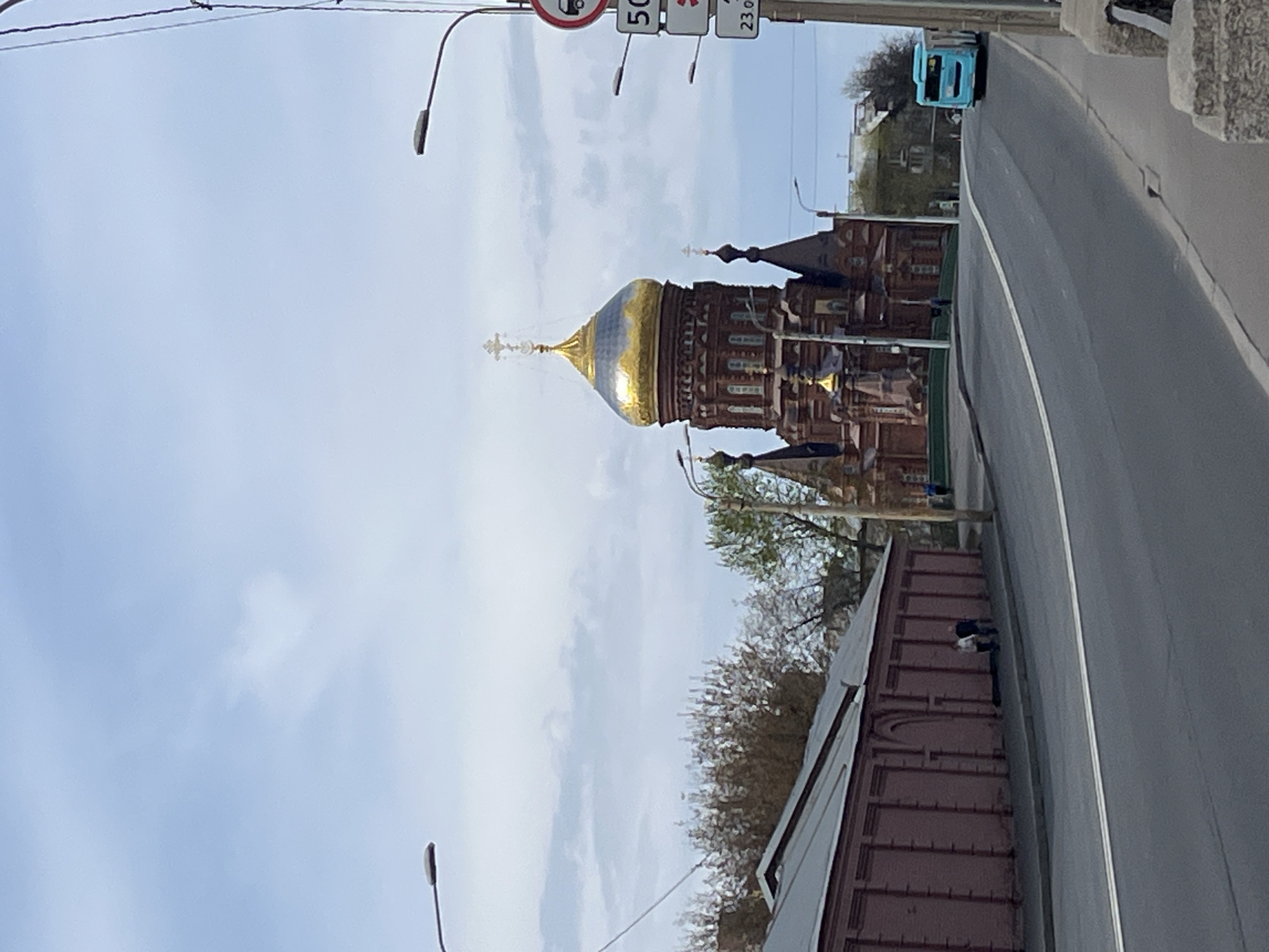 The image shows a view in what appears to be Saint Petersburg, Russia, looking towards a domed cathedral. The cathedral has a large, golden dome with a cross on top. The architecture of the building is characterized by reddish-brown brick. Alongside the road to the right of the cathedral, there is a truck driving. Traffic signs are visible above the truck. The sky is overcast.