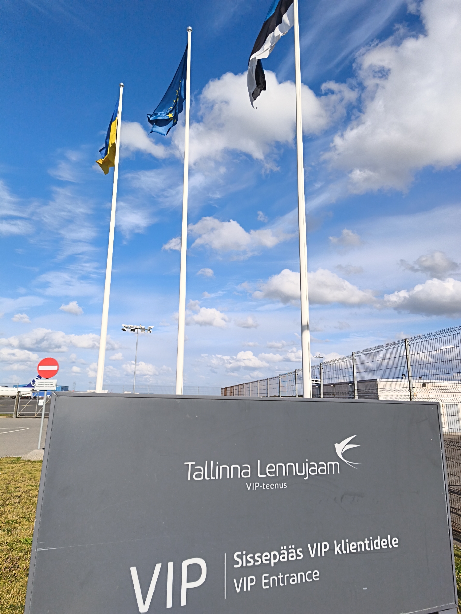 The image captures a scene outside Tallinn Airport in Estonia on a bright, cloudy day. Three flagpoles stand tall, flying the flags of Ukraine, the European Union, and Estonia. In the foreground, a large, dark grey sign prominently displays "Tallinna Lennujaam" (Tallinn Airport) along with the text "VIP-teenus" (VIP service) and "Sissepääs VIP klientidele" (Entrance for VIP clients) and "VIP Entrance". The sign has a stylized bird logo. To the left of the sign, a red circular sign with a white horizontal bar indicates a no entry or restricted area. In the background, a chain-link fence encloses an area with some buildings, and a white airplane is visible in the distance. The overall environment suggests an airport tarmac or a service area within the airport grounds.