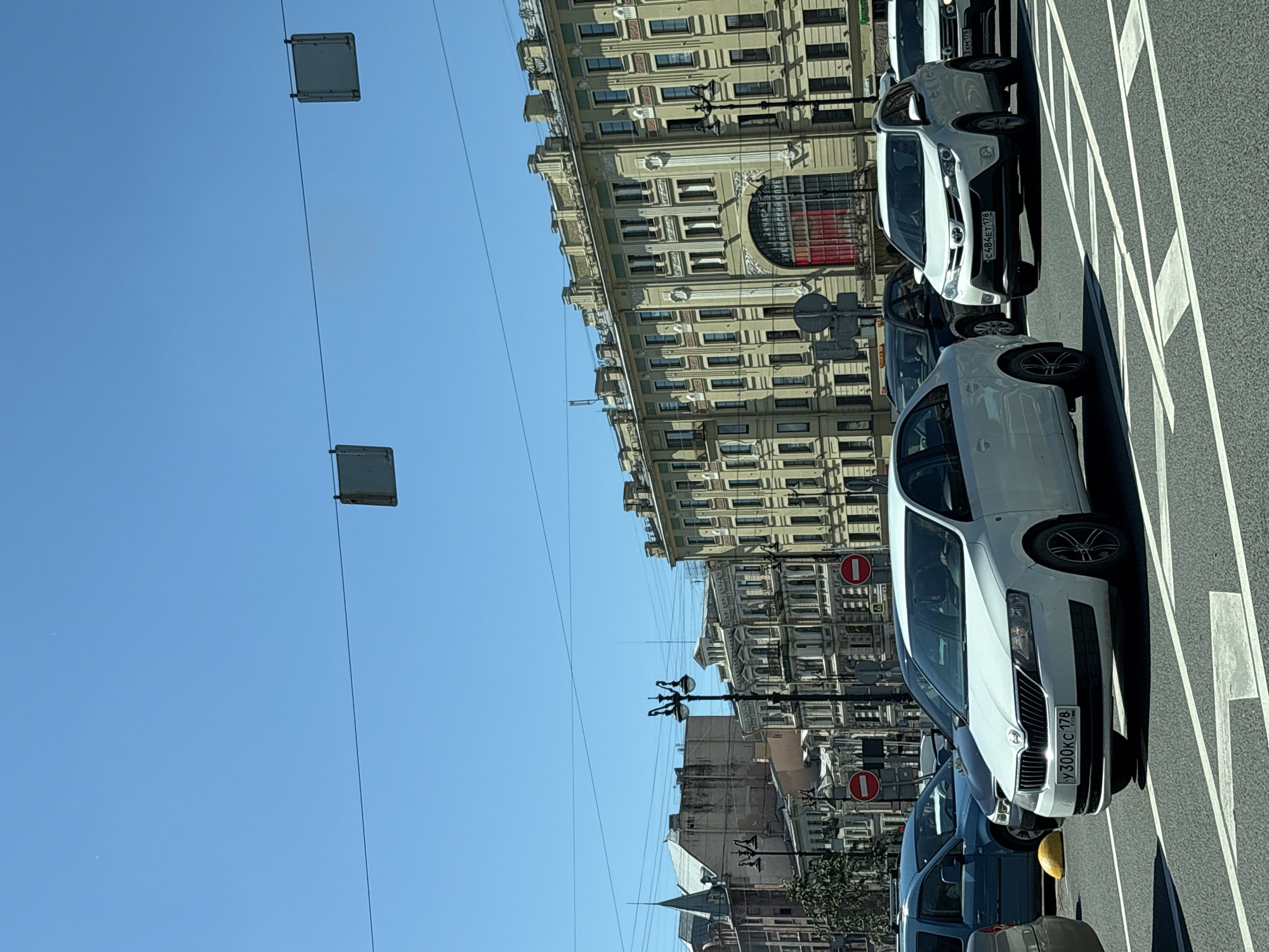 This is a daytime, eye-level, medium shot of a city street in Saint Petersburg, Russia, depicting a traffic intersection under a clear blue sky. The foreground is dominated by several cars, including a white Skoda Rapid with a Russian license plate that reads "У300КС 178". To its right is a white Toyota RAV4 with a license plate "C484ET 178". Behind these vehicles, a blue car and a black car are visible.
The background features a row of ornate, multi-story buildings with yellow facades, characteristic of Saint Petersburg's architecture. These buildings have numerous windows and decorative elements, including arches and columns. Street lamps with multiple arms are visible along the sidewalk. Traffic signs, specifically circular "no entry" signs, are positioned at the intersection. Overhead, power lines and square traffic signal boxes are strung across the sky. The road surface is asphalt, marked with white lane lines, indicating a complex intersection. The overall scene conveys a busy urban environment with ongoing traffic flow.