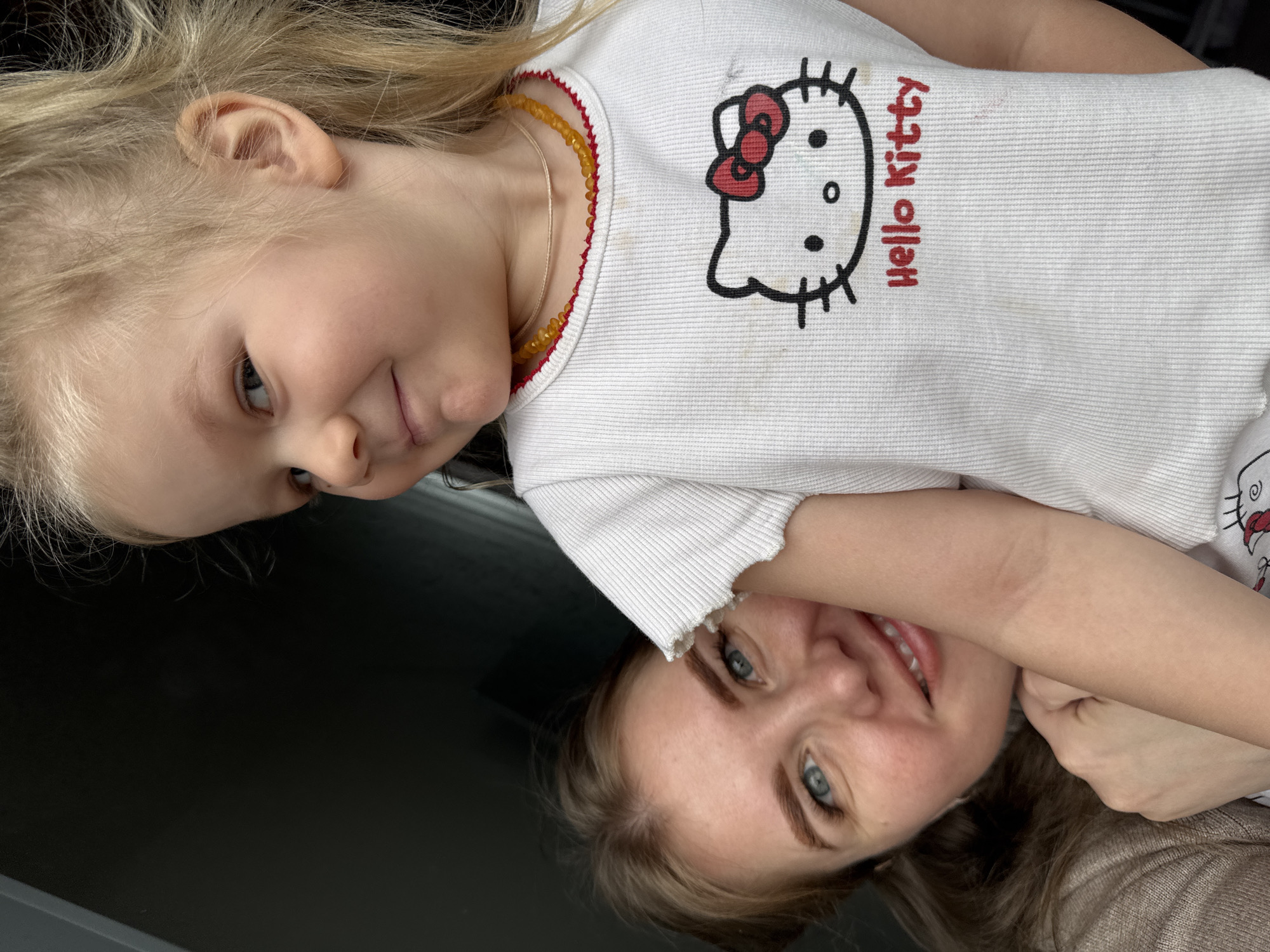 A young girl with blonde hair and a "Hello Kitty" t-shirt is smiling. An adult woman, possibly the girl's mother, with light blonde hair and blue eyes, is smiling and holding the girl close. They appear to be indoors, possibly in a house, based on the lighting and visible surfaces. The location is Gornyi (Dvurechenskoe S/P), Russia, as per the prompt's instructions, though this cannot be directly determined from the image.