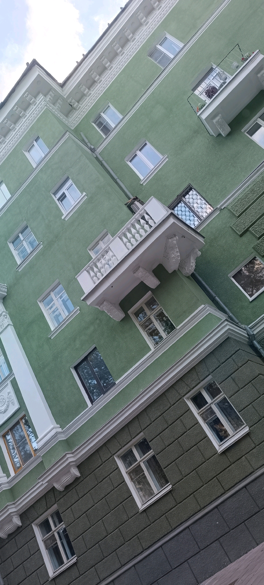 The image shows the exterior of a green, multi-story building, likely an apartment building, viewed from a low angle looking upwards. The architecture features ornate white trim along the roofline and around windows. There are several windows visible, some with open shutters or balconies. One balcony extends from the building, supported by decorative brackets, and has potted plants on it. The building's facade has a textured finish. The lower section of the building appears to be a different color, a darker green with a brick-like pattern. The image is tilted, suggesting it was taken with the camera at an angle. The scene is likely located in the city of Perm, Russia, given the context of previous similar images. There are no people visible in the image.