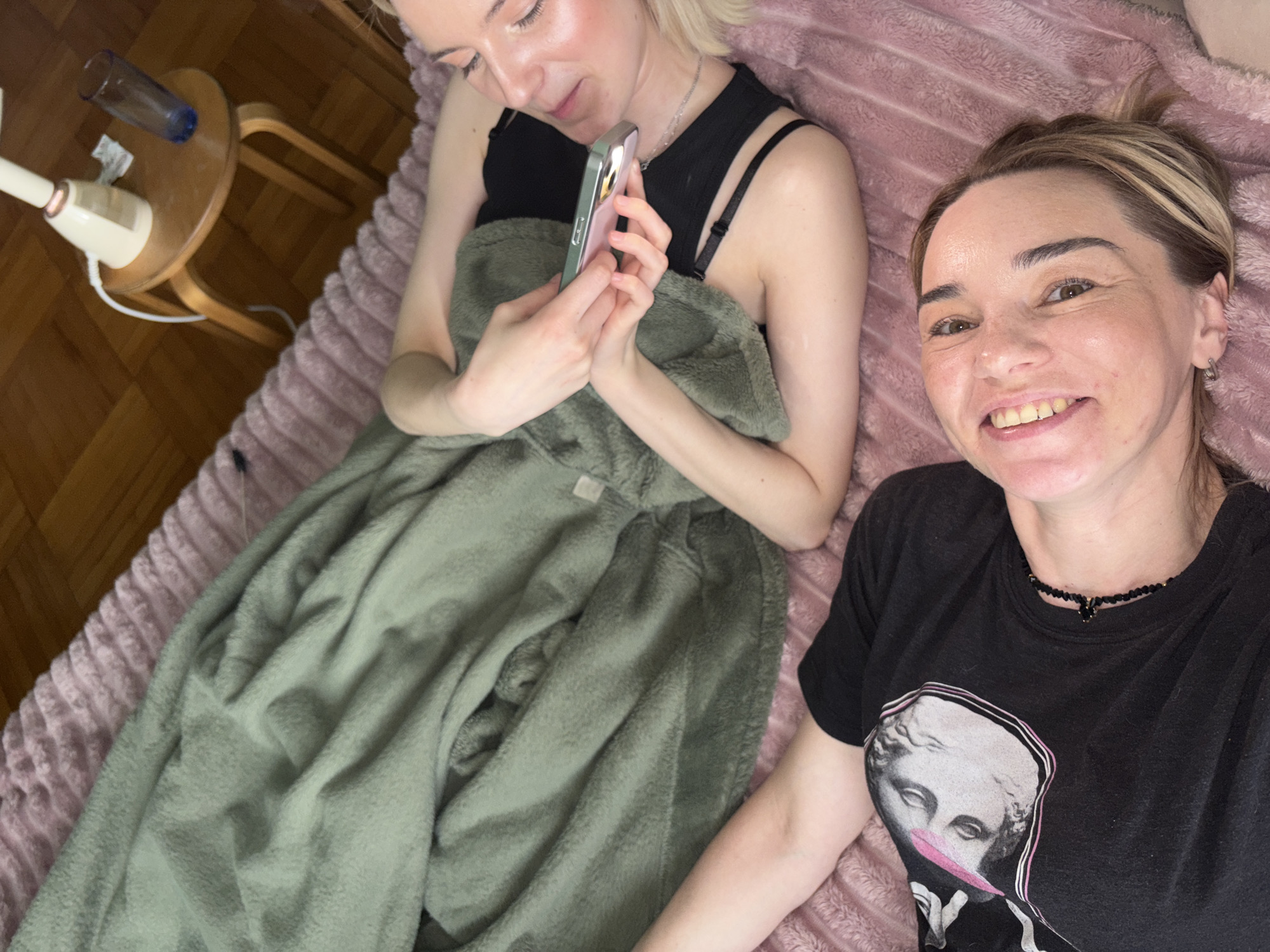 Two women are captured in a relaxed, overhead shot, likely indoors in Vsevolozhsk, Russia. They appear to be lying on a bed or sofa covered by a thick, textured pink blanket.
The woman on the left, with blonde hair and fair skin, is seen in profile looking down at a light-colored smartphone with a pink case, which she holds with both hands. She is partially covered by a soft, olive-green fuzzy blanket or robe. She wears a black top with visible straps. Her expression is calm and focused on her device.
The woman on the right, with brown hair and blonde highlights, is smiling directly at the camera. She has a warm and happy expression. She wears a black t-shirt featuring a graphic print of a classical bust with a modern pink overlay. A dark beaded necklace is visible around her neck.
To the left of the blonde woman, a light-colored wooden side table is visible on a wooden parquet floor. On the table, there's an inverted blue glass, a crumpled white tissue or paper, and a white and beige electric device, possibly a humidifier or diffuser, with a white cord extending from it. The overall setting suggests a cozy and intimate atmosphere, possibly in a bedroom or living area. No specific time of day or weather cues are evident. There is no clearly legible text visible within the image.