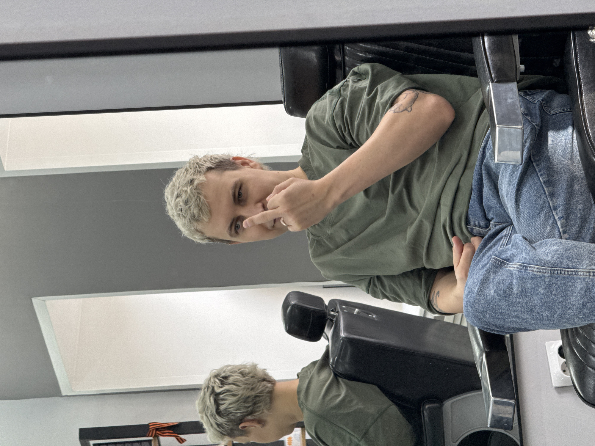 An individual with short, light blonde hair is seated in a black leather barber chair with chrome armrests. The person is wearing a loose olive green long-sleeved shirt and light blue denim jeans. A tattoo, possibly depicting a winged insect, is visible on their left forearm. A gold-colored ring is worn on the left ring finger. The individual looks directly forward, with their right index finger touching their upper lip. A reflection of the individual's back and the barber chair is visible in a large mirror to their left. The background consists of walls painted light gray on the upper section and white on the lower section. A white electrical outlet with two circular sockets is visible on the lower wall. To the far left, a vertical black object is partially visible, adorned with an orange and black St. George's ribbon. The location is an indoor commercial establishment, consistent with a barbershop or salon, in Votkinsk, Russia.