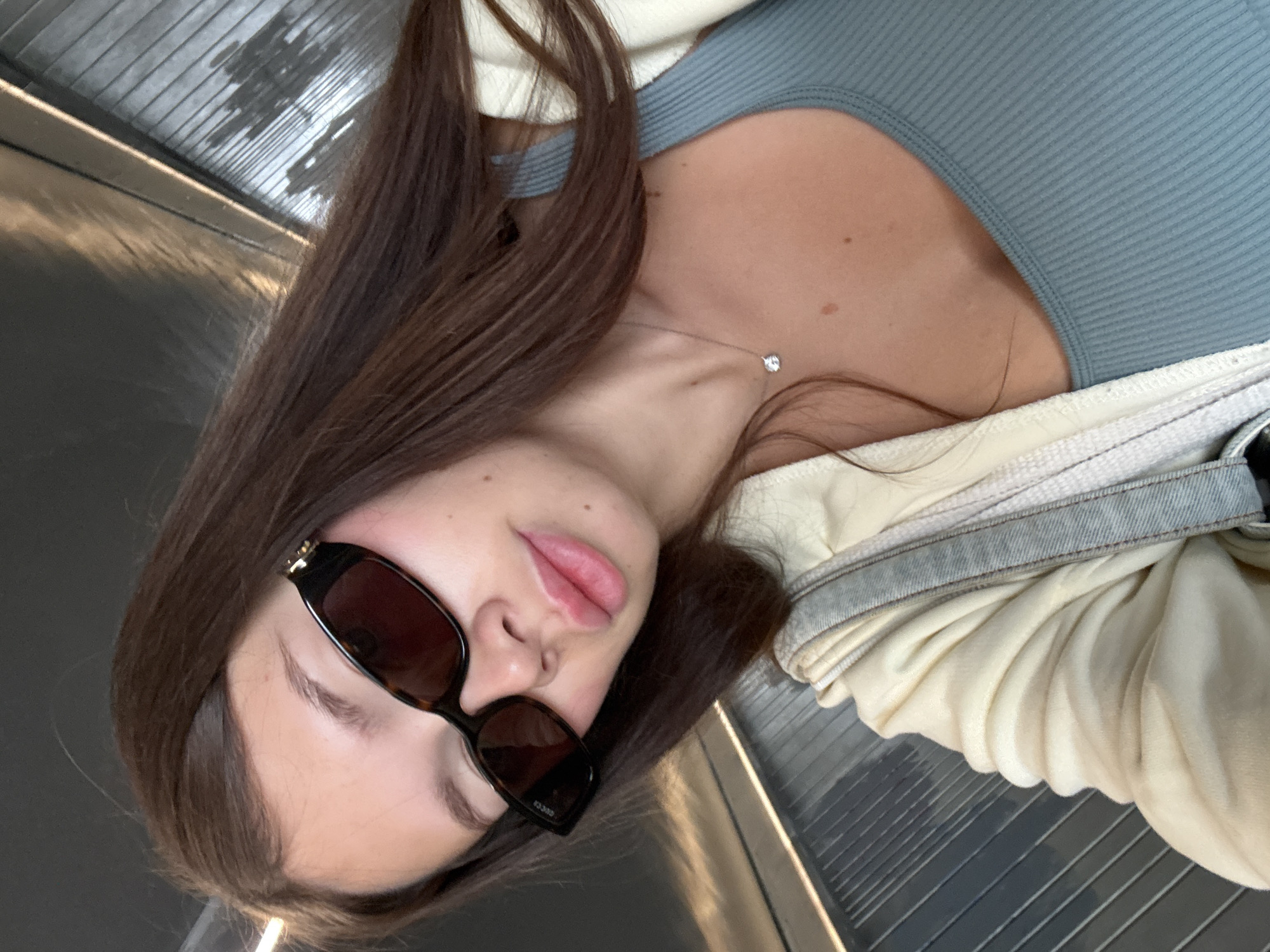 A young woman is captured in a close-up portrait, likely a selfie, rotated 90 degrees clockwise. She has long, dark brown hair flowing around her shoulders and chest. Her face is partially obscured by dark, rectangular sunglasses. Her lips are full and slightly parted in a soft pout, and a mole is visible near her nose/upper cheek. She wears a light blue or teal ribbed top and an off-white or cream-colored, soft jacket, possibly fleece-lined, with a light blue denim-like trim on the sleeve. A delicate, silver-colored necklace with a small, sparkling pendant hangs around her neck. The brand name "GUCCI" is visible on the inner temple of her sunglasses.
The background is dark and features linear patterns on what appears to be a ceiling and a ridged floor, suggesting an indoor environment such as a modern building, a hallway, or perhaps a transportation setting like a train or escalator. There are some indistinct reflections or light patterns on the ceiling. The woman's pose and expression convey a casual demeanor. No specific time of day or weather is discernible from the indoor setting. The location context indicates Tel-Aviv, Israel.