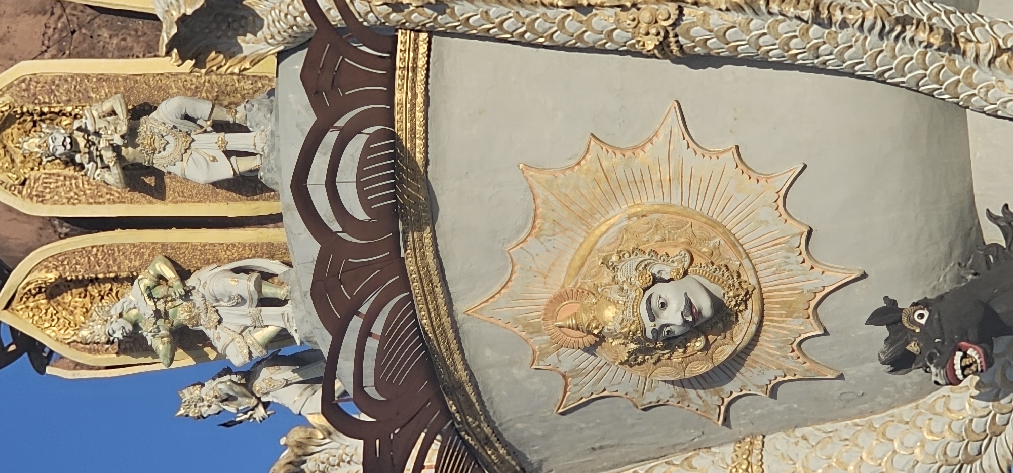 The image captures a detailed architectural feature likely found in Denpasar, Indonesia, given the style of ornamentation. The shot is taken at an upward angle, focusing on a section of a building or monument adorned with religious and decorative elements.
The dominant element is a circular bas-relief featuring a human-like face, possibly a deity or important figure, set within a sunburst-like array of spikes or rays. The face is painted in lighter tones, contrasting with the golden and earthy hues of the surrounding embellishments.
To the left of the central element are tiers of sculpted figures, seemingly statues of deities or mythological beings. They are painted in light shades with gold accents, laid in recessed panels.
On the right side, a dark sculpture of an animalistic figure, possibly a dragon or mythical beast, is visible. It has sharp teeth and pronounced features, adding a contrasting element to the more serene facial representation.
The background is a neutral tone, serving as a canvas for the various decorations. The top of the frame shows more ornate details in gold and white, possibly parts of the roof or upper structure of the building.