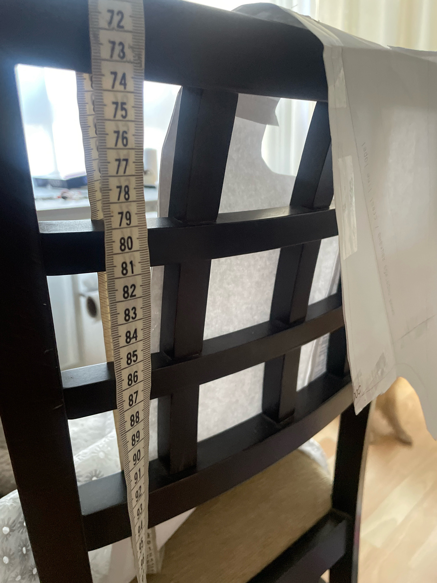 The image shows a measuring tape hanging from a dark wooden chair. The measuring tape is yellow and white with black markings and numbers indicating inches, starting from the top with '72' and going down to '95'. The chair has a slatted back made of dark wood. A piece of white paper or fabric is also attached to the chair. The background is blurred, but it appears to be an indoor setting with natural light coming from a window. There is no identifiable information within the image that suggests a specific location in Atarfe, Spain.