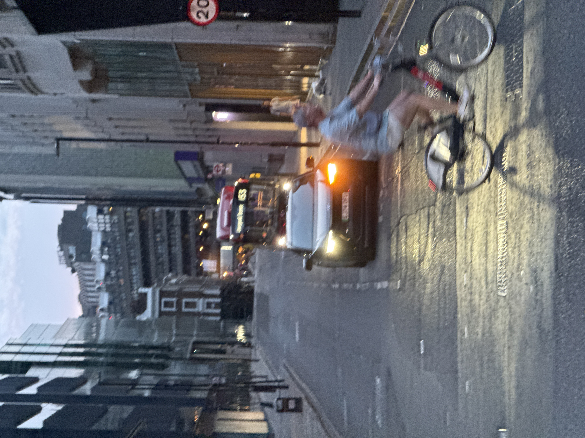 The image shows a street scene in London, United Kingdom. In the foreground, a cyclist rides a bike. A dark-colored car is in the street with headlights on. A red double-decker bus is visible further down the street, along with other traffic. Buildings line the street. A "20" speed limit sign is present.