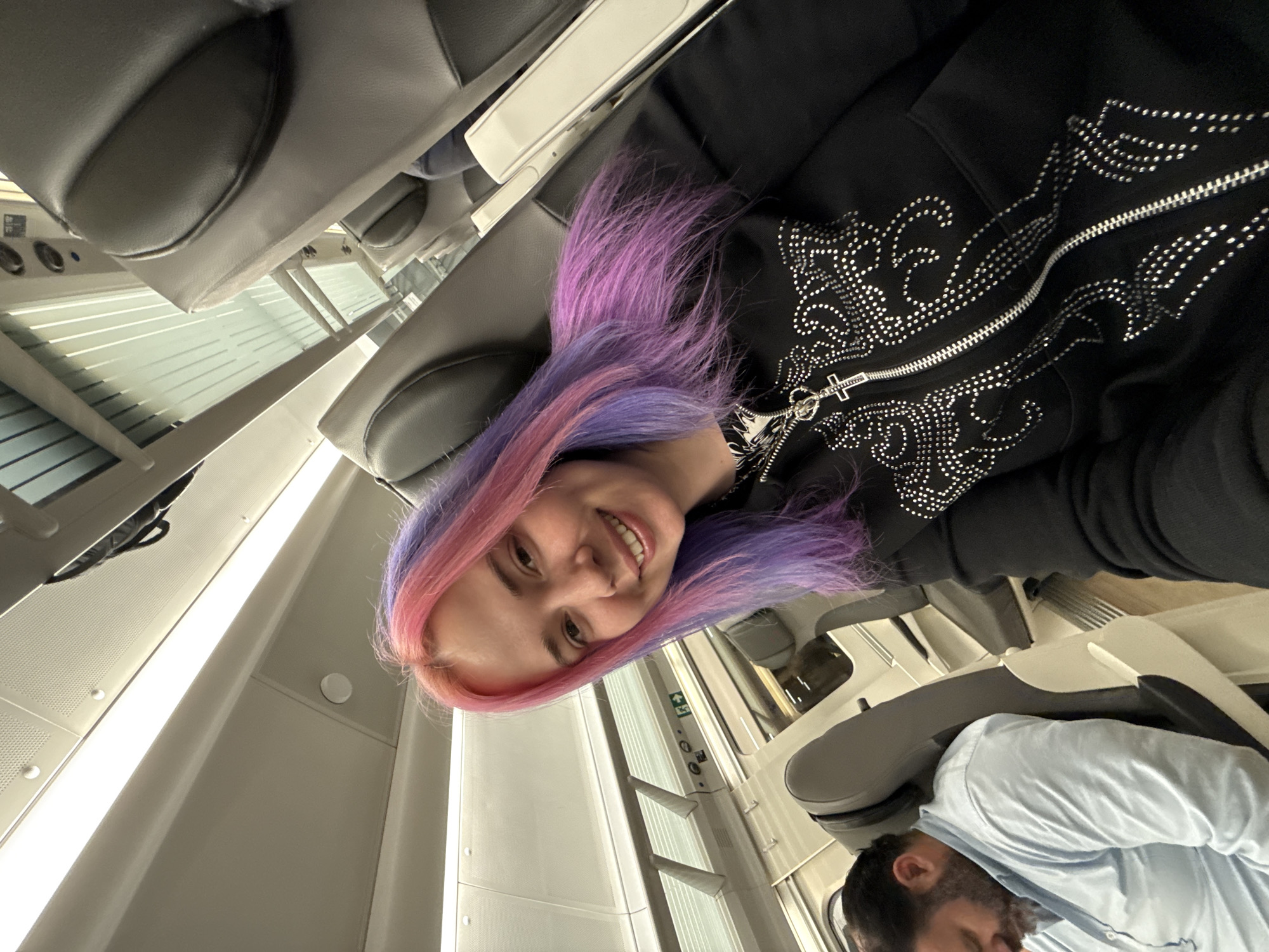 A woman with dyed pink and purple hair is smiling while lying in what appears to be a high-speed train, possibly the AVE in Madrid, Spain, judging from the train's interior. She is wearing a black hoodie with a rhinestone design. A person wearing a blue shirt is visible in the lower right corner of the image. The train cabin has grey seats, white walls, and window blinds.