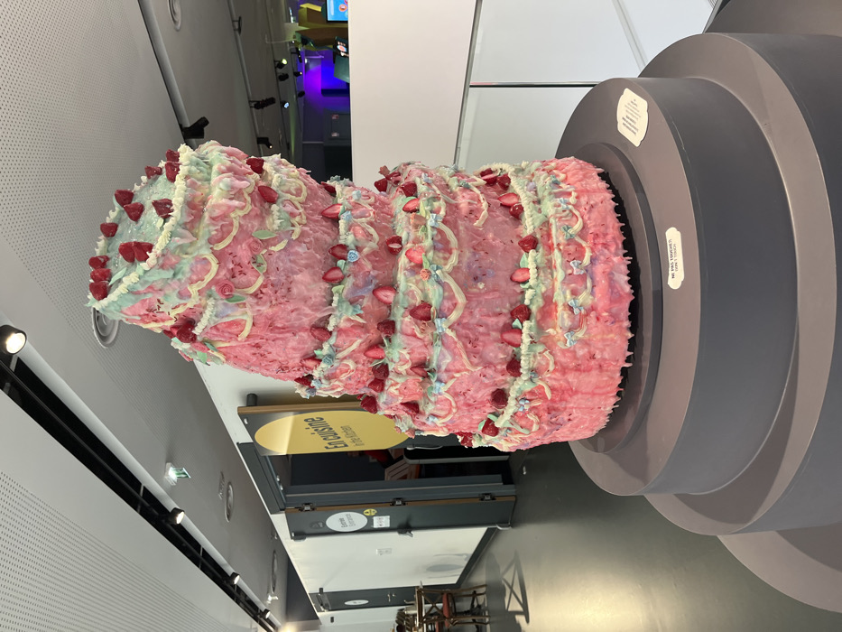 A large, multi-tiered sculpture resembling a melting cake, predominantly pink with dripping white and light blue frosting, adorned with numerous red fruit representations and small blue bows, is displayed on a tiered, dark grey cylindrical pedestal. A white label on the pedestal states "NE PAS TOUCHER / DON'T TOUCH." The sculpture is situated within an indoor exhibition space characterized by a dark floor, white walls, and a white perforated ceiling fitted with black track lighting. In the background, a doorway features a prominent yellow elliptical sign reading "En cuisine / In the Kitchen." Further down the hallway, another door is labeled "Entrée / Entrance." To the far left, a few chairs and tables are partially visible. To the right of the cake, a different exhibit area with distinctive purple and blue lighting is visible beyond a white partition. This scene is located in the Cité de la Gastronomie et du Vin in Dijon, France. No people are visible.
