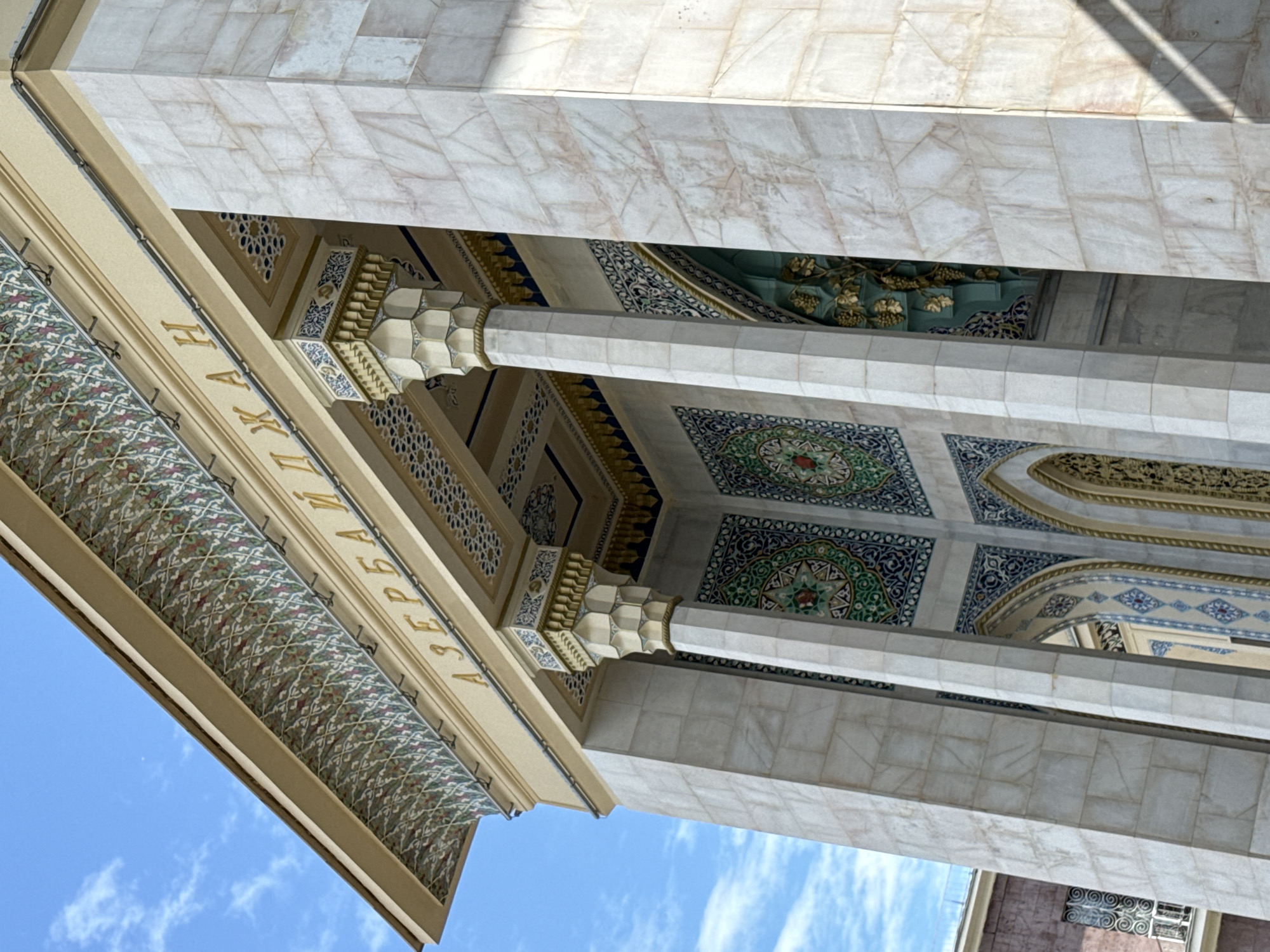 The media file captures an upward-angle view of the ornate facade of a building, likely a mosque or a culturally significant structure, with a clear blue sky and scattered white clouds as the backdrop. The architecture features intricate tilework in shades of green, blue, and white, arranged in geometric and floral patterns. Large, octagonal columns, adorned with decorative elements, support a richly decorated ceiling. The ceiling itself is a mosaic of colorful tiles and gilded carvings.
Golden Cyrillic lettering is visible along an architectural beam, partially obscured by the angle and focus. The text appears to read "АЗЕРБАЙДЖАН" (Azerbaijan). The building's exterior also showcases large marble-like panels and a patterned awning or canopy. The lighting suggests it is daytime, likely mid-morning or afternoon, with sunlight casting shadows on the marble surfaces. The overall impression is one of grandeur and cultural richness, typical of Islamic architectural styles. No people are visible in the frame. The scene is static, with no discernible activity other than the architectural display under a clear sky. The location context provided indicates Moscow, Russia.