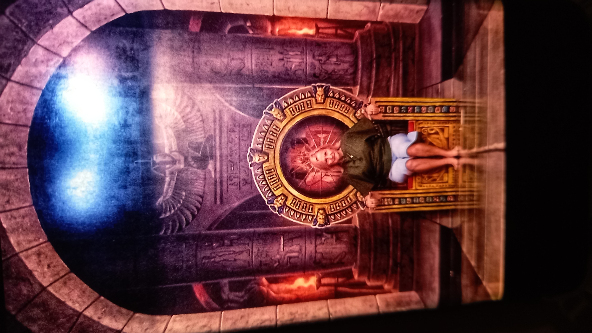 A woman sits on a throne in front of an Egyptian-themed mural. She is wearing a green top and blue shorts, and her feet are bare. The throne is ornate, with gold accents and carvings. The mural depicts a winged figure, hieroglyphs, and columns. The overall scene appears to be in a dimly lit room, possibly a tourist attraction or themed exhibit. The location is not specified as being in the city of ตำบลเกาะพะงัน, Thailand.