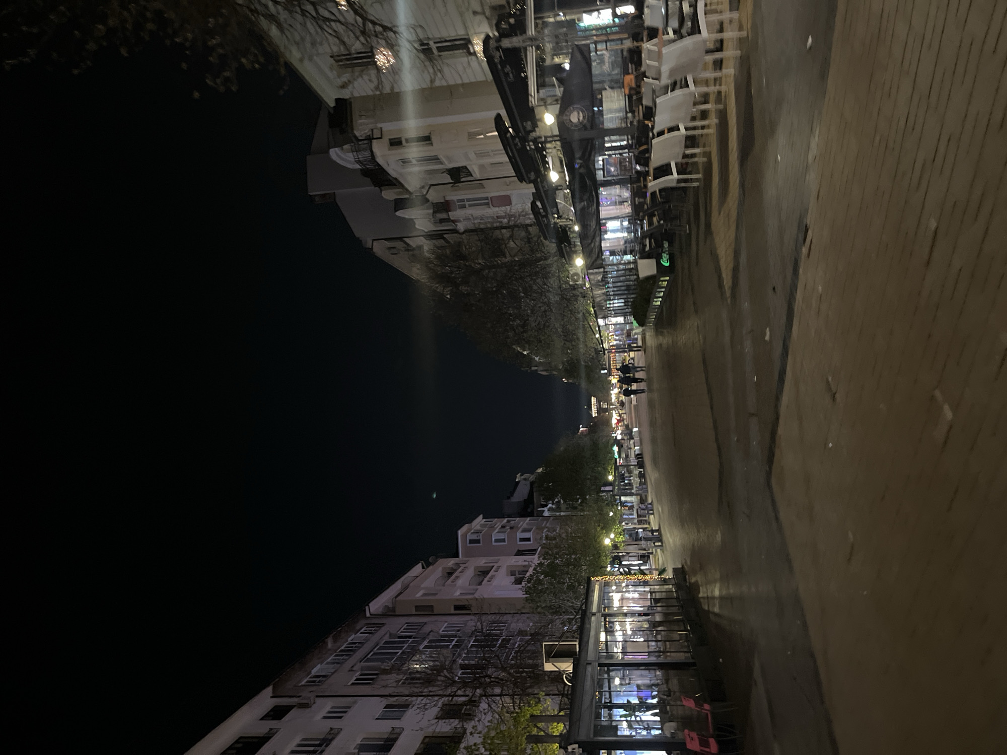 The image depicts a night scene on a street in Sofia, Bulgaria. Buildings line both sides of the street, with multi-story apartment buildings visible on one side and commercial establishments with outdoor seating on the other. The sidewalk is paved. Trees are present, adding to the urban landscape. Pedestrians are seen walking in the distance. Streetlights illuminate the scene, casting shadows and reflections. Chairs and tables are arranged outside the establishments, suggesting a lively atmosphere. A dark night sky is visible.