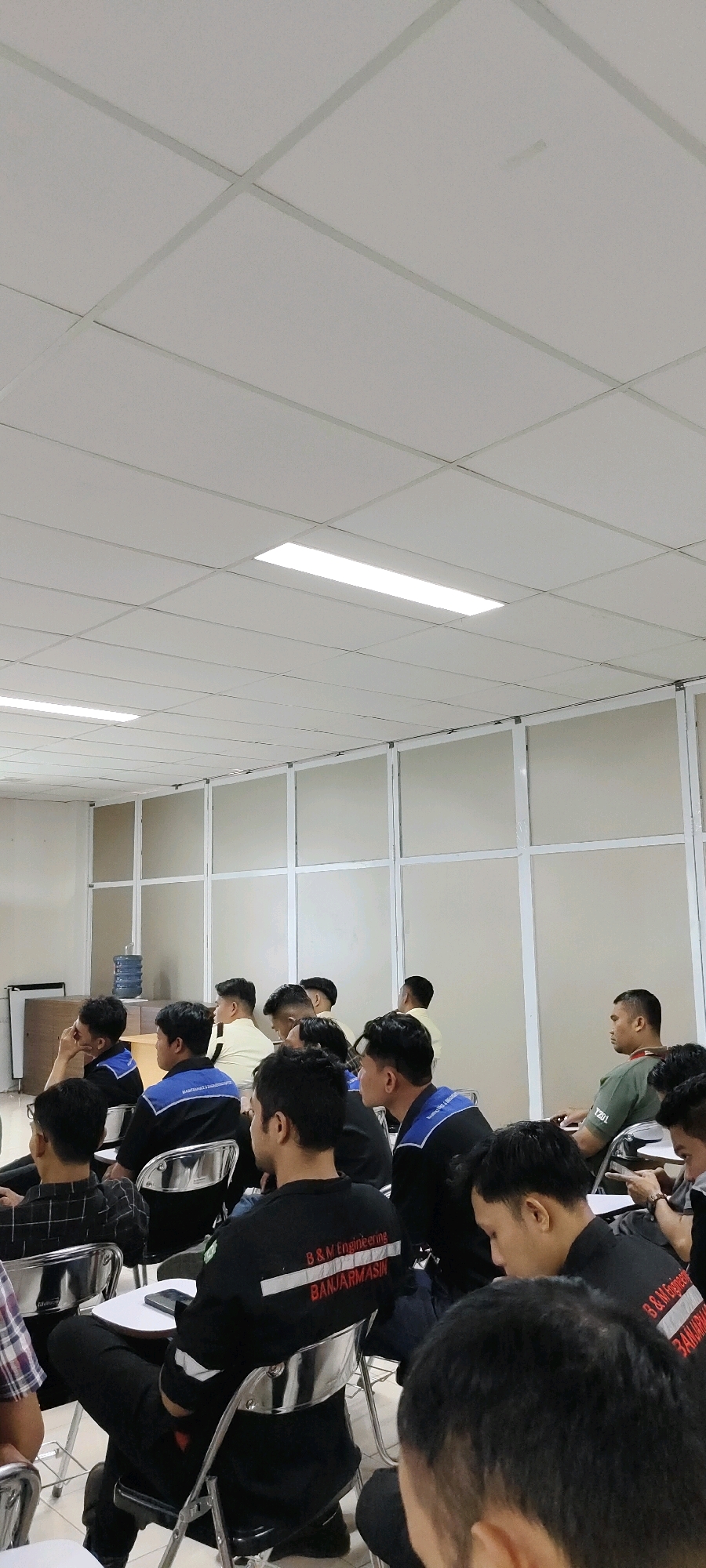 The image depicts an indoor scene, likely a classroom or lecture hall, where a group of young men are seated, facing forward. The ceiling is a grid of white panels with fluorescent lights. Partitions with frosted glass are visible along the back wall, separating the space. In the foreground and mid-ground, the men are predominantly wearing black shirts, some with blue accents and white lettering that reads "B&M Engineering BANJARMASIN". They are seated on metallic folding chairs, with some chairs having small white tables attached. A water cooler is visible to the left, behind a desk. The lighting is even and bright. The individuals appear to be attentive and engaged in whatever is happening in front of them. The presence of the "BANJARMASIN" text on their clothing suggests a connection to that city. This scene is consistent with an educational or training session.