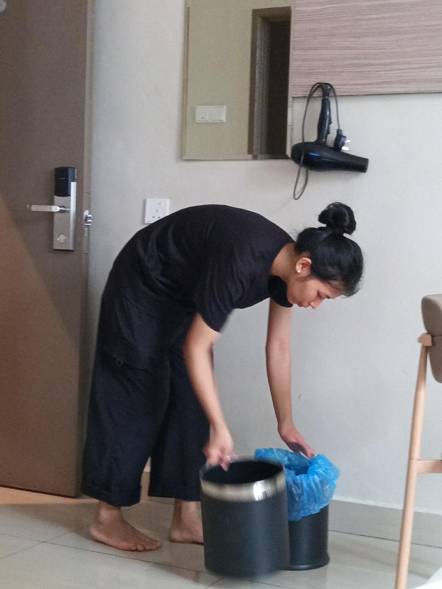 In Kuala Lumpur, Malaysia, a woman with her hair in a bun, wearing a black t-shirt and wide-leg black pants, is bending over to empty a black trash can with a metallic rim into another black trash can lined with a blue plastic bag. She is barefoot and standing on a tiled floor. To her left, a brown door with a silver electronic lock is visible. In the background, a wall-mounted hairdryer hangs on a wooden panel.