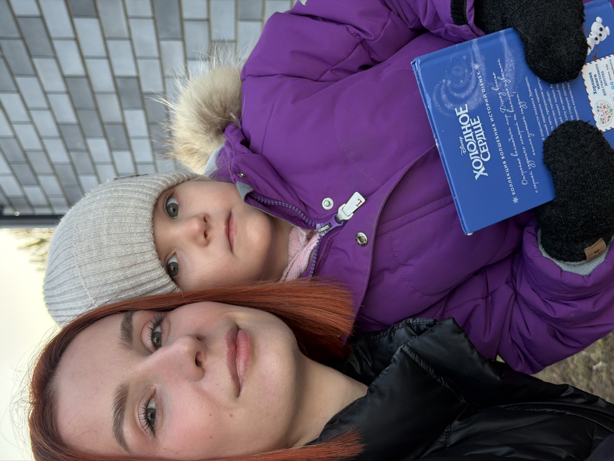 In Chaikovsky, Russia, an outdoor close-up image depicts an adult female and a young female child. The adult, positioned on the left, has fair skin, auburn shoulder-length hair, and light eyes, with a slight smile as she looks towards the camera. She is wearing a black puffer jacket. To her right, the young child wears a light grey knit beanie, a purple puffer jacket with a fur-lined hood, and black knitted gloves. The child's light eyes are open, and she holds a blue book titled "ХОЛОДНОЕ СЕРДЦЕ" (FROZEN) with the subtitle "КОЛЛЕКЦИЯ ВОЛШЕБНЫХ ИСТОРИЙ DISNEY" (Collection of Magical Disney Stories) visible on its cover. Both individuals face the camera. The background consists of a grey brick wall behind the child, with a clear sky visible above it. Blurred brownish-green ground is visible in the lower left corner.
