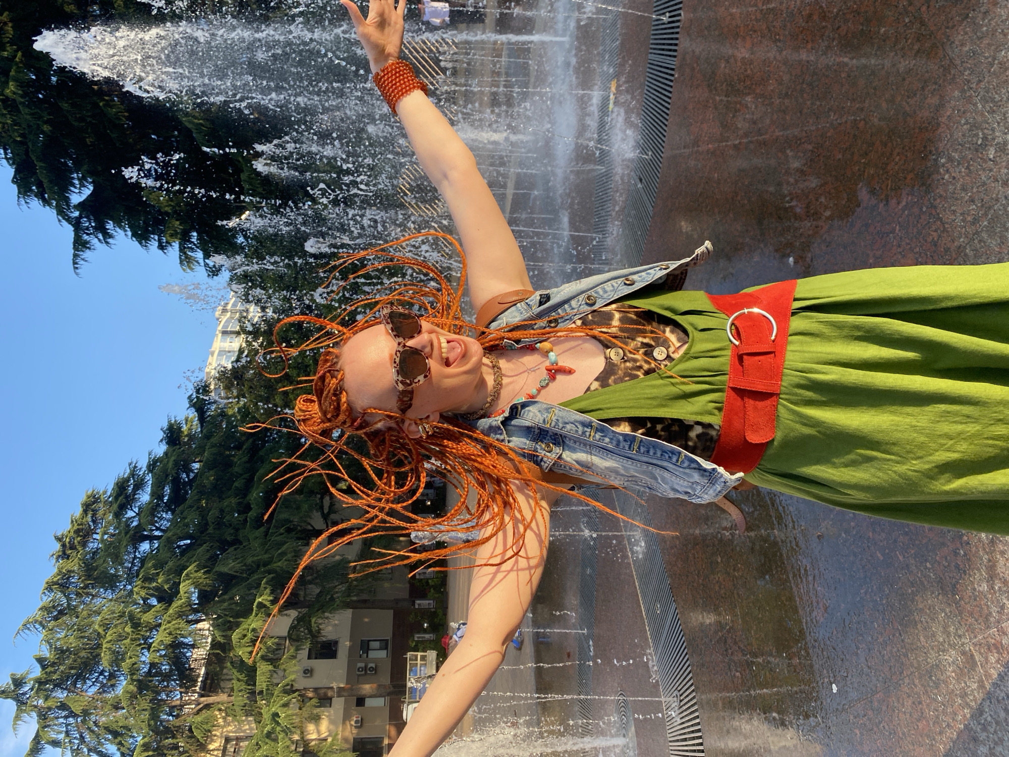 A woman with bright orange braided hair is joyfully posing with her arms outstretched in front of a water fountain in Sochi, Russia. She is wearing a green dress with a red belt, a denim vest, and sunglasses. The fountain sprays water high into the air, creating a refreshing mist. Tall green trees line the background, and a building is visible behind them. The sun is shining, casting a warm glow on the scene.