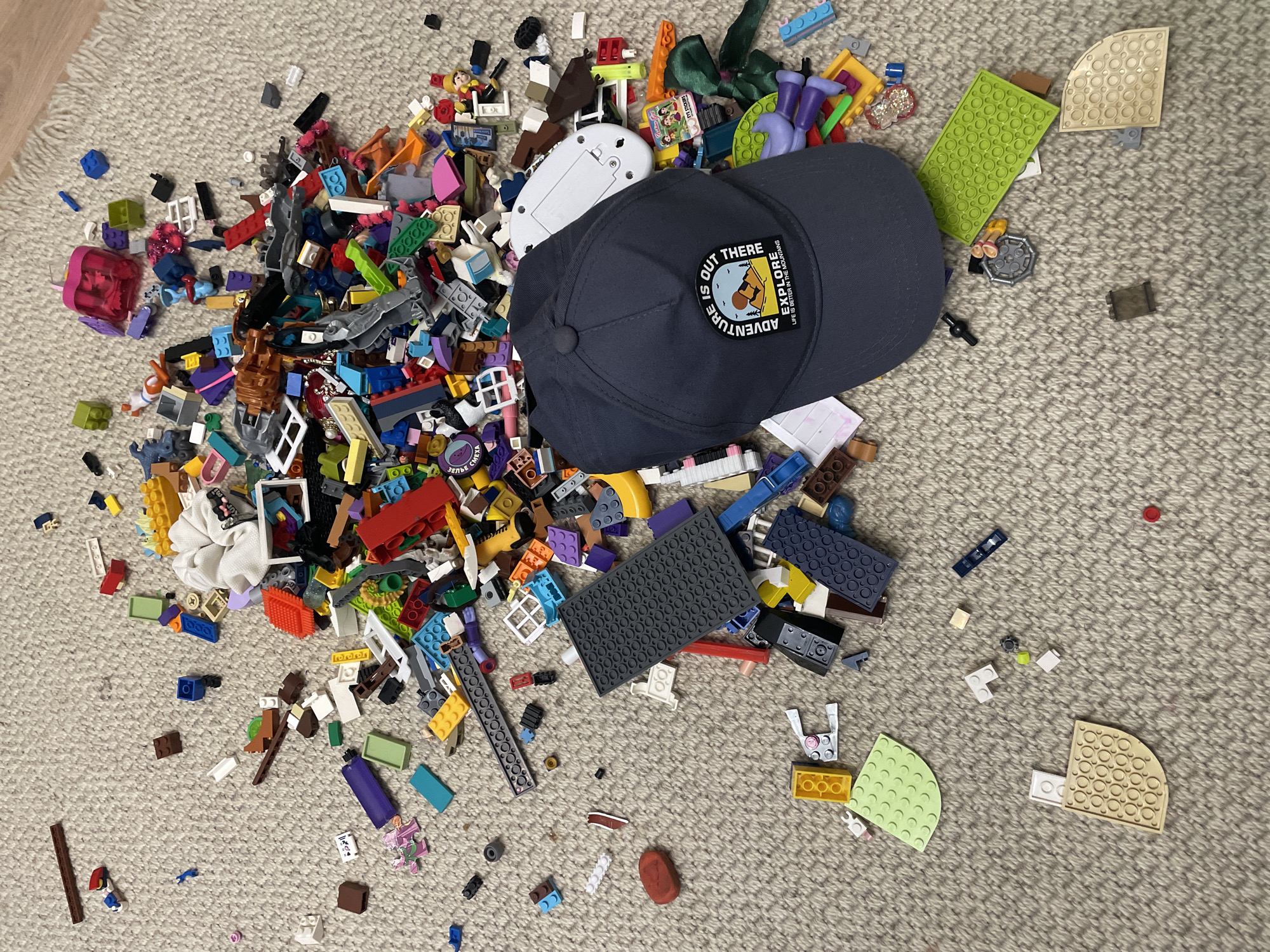 A dark blue baseball cap with a circular patch is placed on a carpeted surface, surrounded by a large assortment of scattered Lego bricks and other small toys. The patch on the cap reads "ADVENTURE IS OUT THERE EXPLORE LIFE IS BETTER IN THE MOUNTAINS". The Lego pieces are of various colors, shapes, and sizes, including larger base plates and smaller individual bricks. Some Lego figures and accessories are also visible. The carpet is a neutral, textured beige. The scene appears to be a typical disarray of children's toys. The image does not contain information that would indicate its location in the city of Cahul, Moldova, or any specific geographical information.