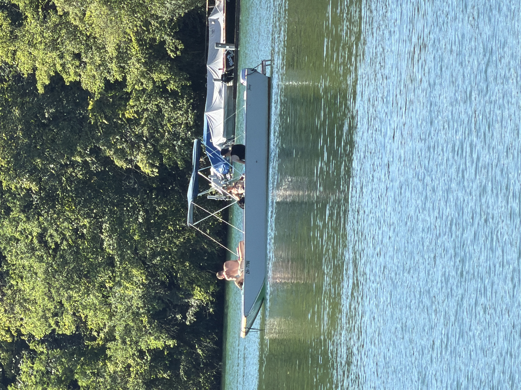 This image captures a serene scene on the Rhine River in Eglisau, Switzerland. In the foreground, the clear, blue water shimmers with ripples. In the middle ground, a long, narrow boat with a light-colored hull is positioned horizontally. The boat has a blue canopy supported by metal poles.
On the bow of the boat, a shirtless person is lying down, facing away from the camera. To their right, another person is seated and appears to be interacting with someone else in the boat. Further back in the boat, another individual is visible, wearing a dark top. The boat has a license plate that reads "ZH-7756."
In the background, a dense line of lush green trees forms a natural backdrop. Behind the boat and further in the distance, there are other docked boats, some covered with white tarpaulins, suggesting a marina or waterfront area. The overall impression is one of a tranquil summer day on the river, with people enjoying leisure time on the water.