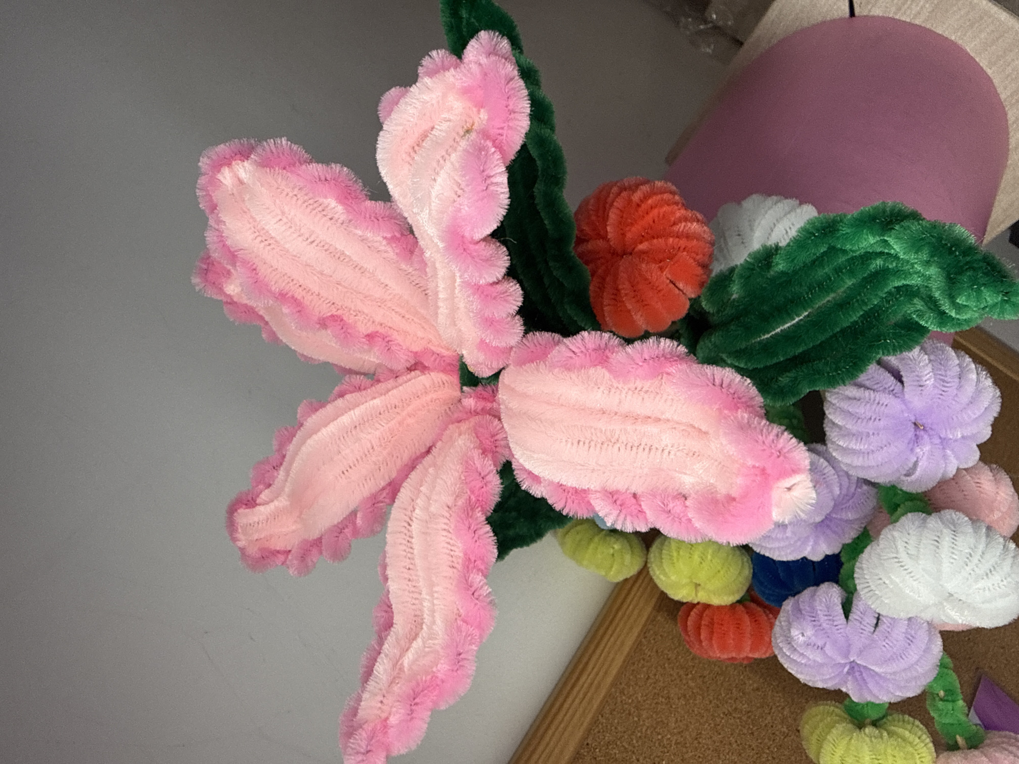 The image displays an arrangement of handcrafted flowers made from pipe cleaners. A large pink flower with frilly edges dominates the composition, accompanied by green pipe cleaner leaves, an orange pipe cleaner flower, and various other pastel-colored pipe cleaner flower heads in shades of white, purple, yellow and pink. The arrangement appears to be placed on a bulletin board or corkboard, as visible from the cork texture in the bottom right corner. A portion of a purple object is visible in the upper right corner. The setting appears to be indoors, likely a craft space or similar environment. The image was taken at an angle looking down on the flower arrangement. The location (Troyan, Bulgaria) cannot be confirmed from the visual information alone.