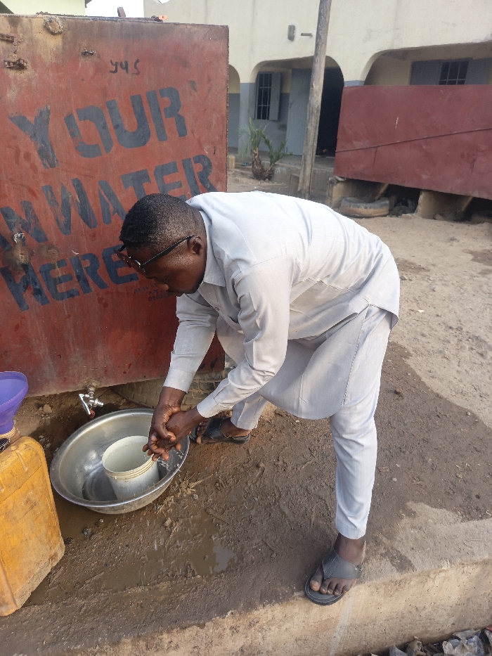 A man washes his hands outdoors at a water station. He wears glasses, a gray shirt and pants, and dark sandals. He leans over a metal bowl under a faucet, with a smaller bucket inside the bowl. A red water tank stands behind him, with white lettering that reads "YOUR WATER HERE." Muddy ground surrounds the station. A yellow container and a purple funnel are visible beside the bowl. In the background, a building with arched windows and a covered porch can be seen.