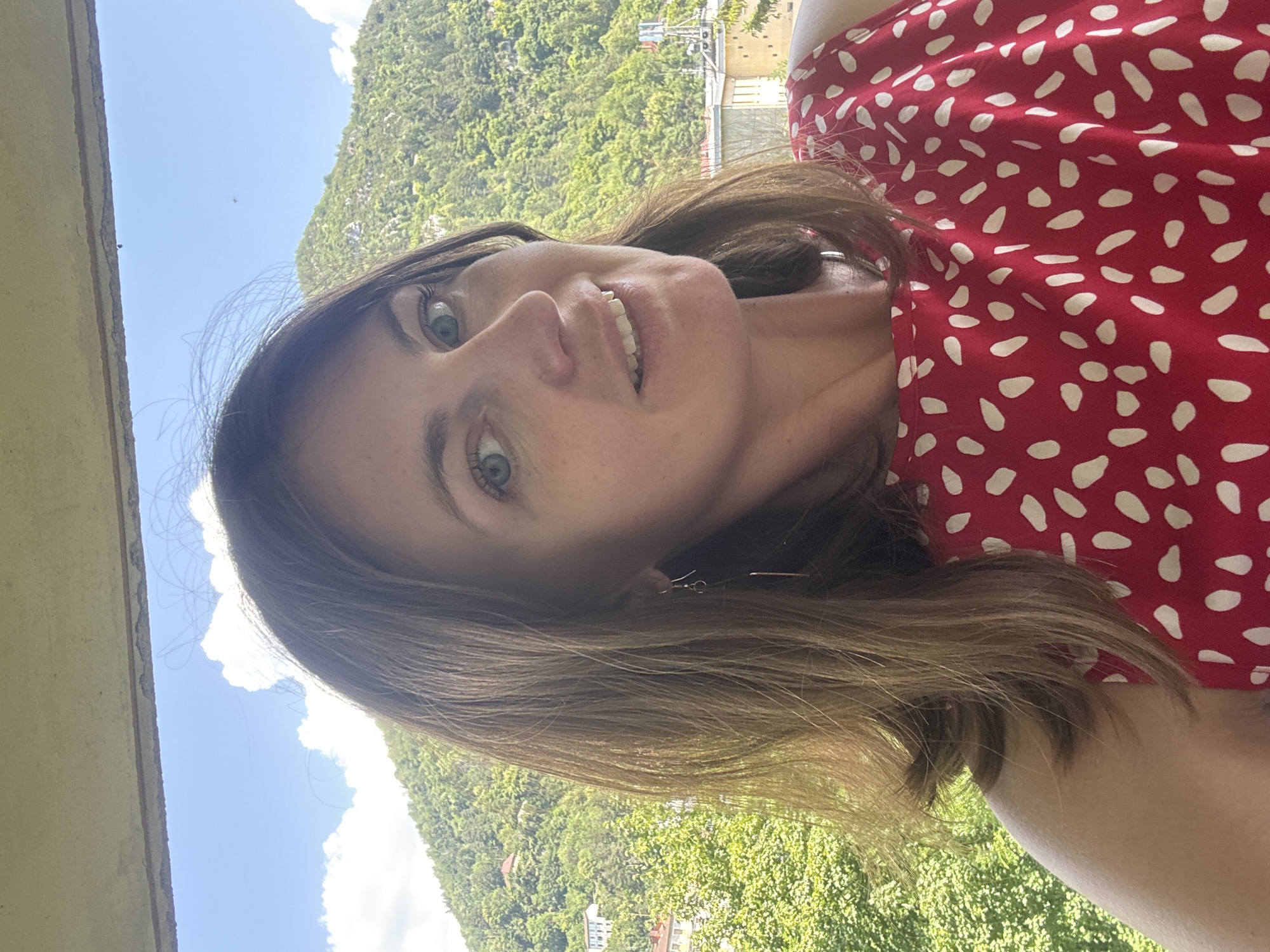 A woman with shoulder-length brown hair and light blue eyes, wearing a red and white polka dot dress, smiles at the camera. She is outdoors, with a lush green mountainside in the background. Buildings are visible on the slopes of the mountain, suggesting an urban or semi-urban environment. The sky above is a clear blue with scattered white clouds. The image appears to be a selfie, taken from a slightly elevated position, possibly from a balcony or porch, overlooking the city of Saint-Claude, France, based on the architecture and mountainous terrain.