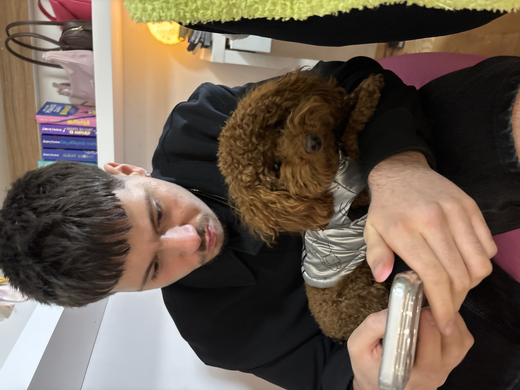 A man with short, dark hair and stubble is holding a small, brown poodle wearing a silver jacket. The man is wearing a black shirt. The background includes a white shelf with books and bags, a yellow light fixture, and a pink cushion. The image appears to be taken indoors. There is no information in the image to place the subject in Sofia, Bulgaria.