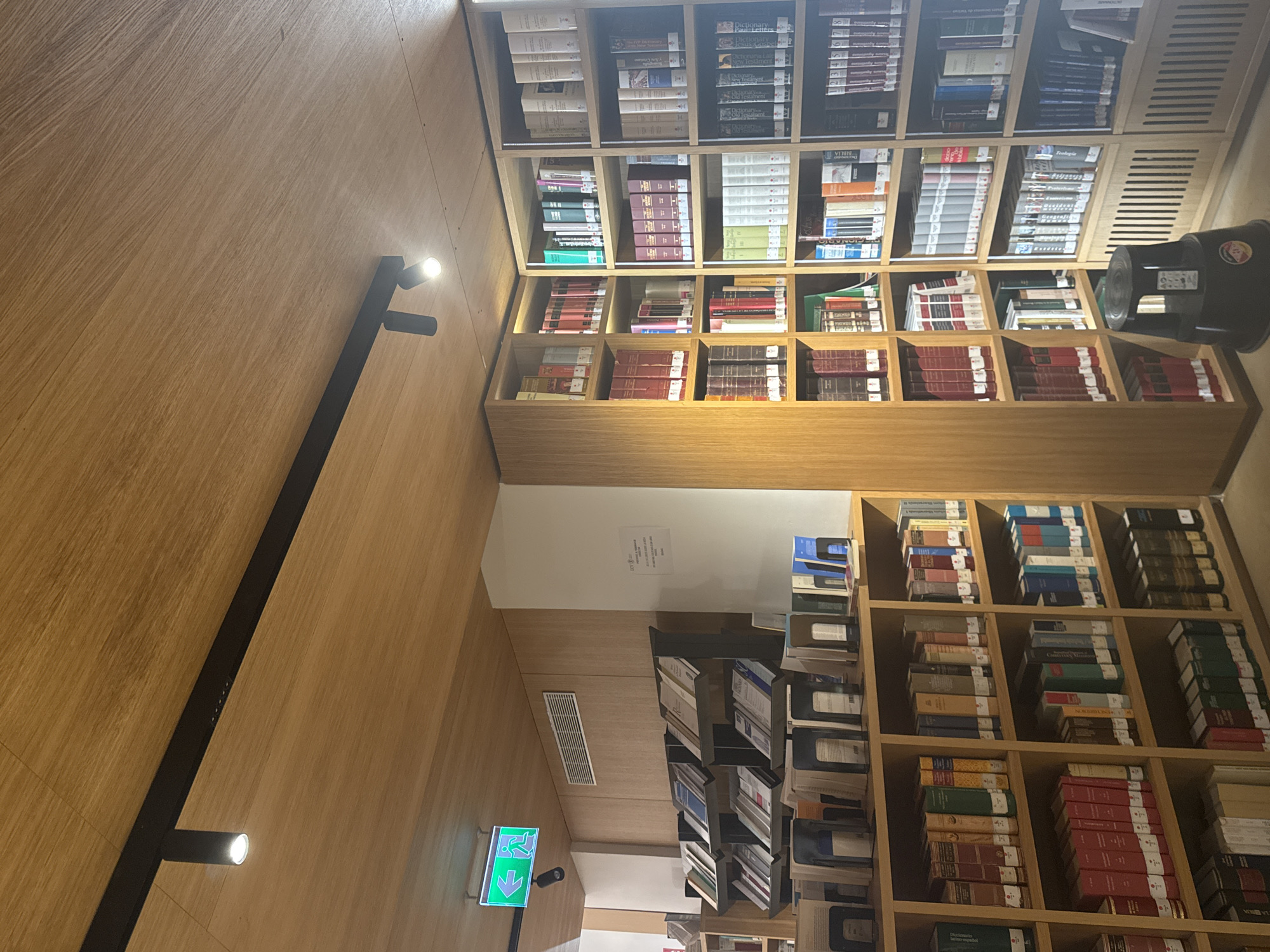 This image displays the interior of the Biblioteca Valenciana (Valencian Library) located in València, Spain. The scene is dominated by extensive wooden bookshelves filled with books, arranged against wooden walls and beneath a wooden panel ceiling.
The ceiling features a black track lighting system with multiple cylindrical spotlights providing illumination. On the left side of the frame, a green illuminated exit sign with a white running figure pointing left is affixed to the wall. Below this, a section of shelving presents books at an upward angle, supported by metal bookends. A rectangular air vent is visible on the wall above this section.
A white vertical pillar located centrally on the right side of the image displays a sign with partially legible text, including "BCV" (Biblioteca de la Comunitat Valenciana), "PERSONAS A INVESTIGAR," "SOLO CON CITA PREVIA," and instructions regarding the "RECOGIDA Y DEVOLUCIÓN DE LIBROS," confirming the library's operational details.
The bookshelves are densely packed with books, many featuring uniform bindings in red, black, and lighter tones, suggesting academic or reference collections. In the bottom right foreground, a black, two-step utility stool with a handle is positioned, and a remote control rests on its top step. The lower sections of some built-in bookshelves incorporate slatted wooden panels. No individuals are present in the visible area of the library.
