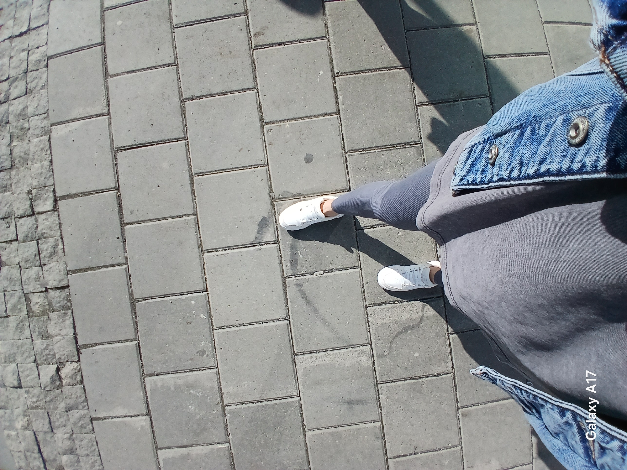The image, taken from a high-angle perspective, shows a person's legs and feet walking on a city sidewalk in Yekaterinburg, Russia. The sidewalk is paved with rectangular gray interlocking concrete blocks. The person is wearing white sneakers, gray leggings, and a gray t-shirt, with a denim jacket draped over their shoulders. A shadow cast by the person is visible on the right side of the frame. The timestamp "Galaxy A17" is visible in the bottom left corner. The lighting suggests it is daytime, with sunlight creating distinct shadows. The overall scene depicts an ordinary urban environment.