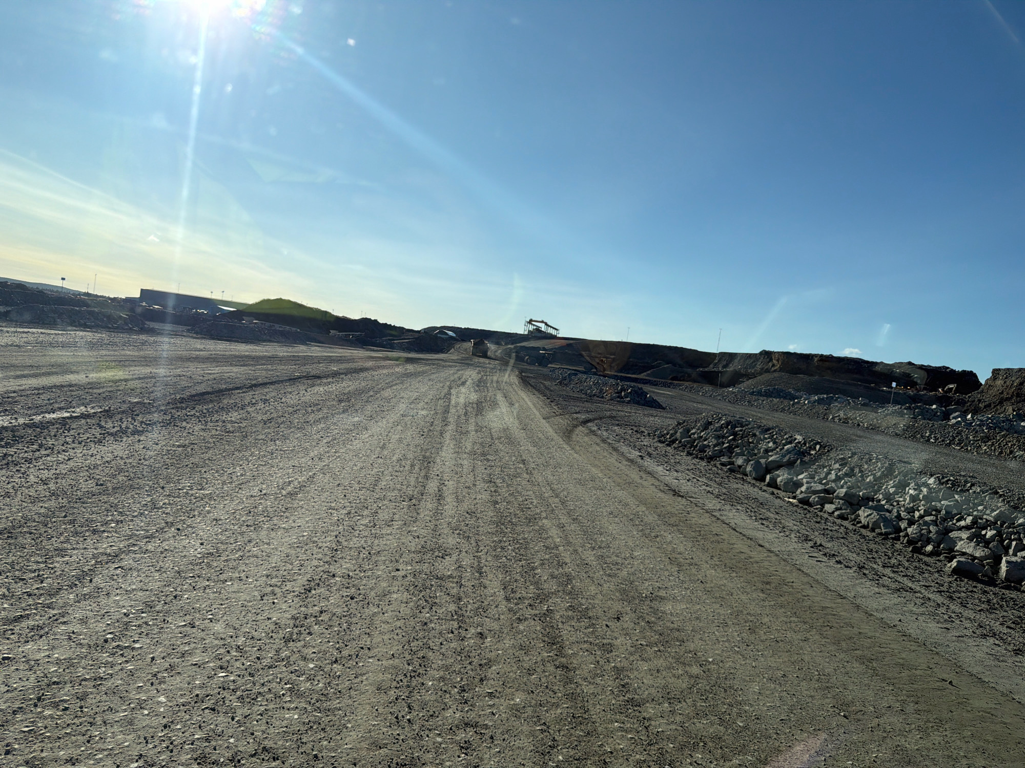 This image depicts an outdoor scene in an industrial area, likely a quarry or mine, in Gällivare, Sweden. A wide, unpaved dirt road dominates the foreground, showing tire tracks from vehicles. The road leads into the distance, curving slightly to the right. On either side of the road are piles of gray gravel and large rocks, indicative of excavation or construction.
In the background, the terrain rises, forming berms and mounds of earth and rock. Several pieces of heavy machinery, including what appear to be excavators or cranes, are visible on the higher ground, suggesting ongoing work. A large, long building with a flat roof is situated on the left side of the horizon, possibly a processing facility or storage area. The sky is clear and bright blue, with the sun shining intensely from the upper left, creating lens flare and strong shadows. The overall environment is barren and industrial, characterized by raw materials and heavy equipment.