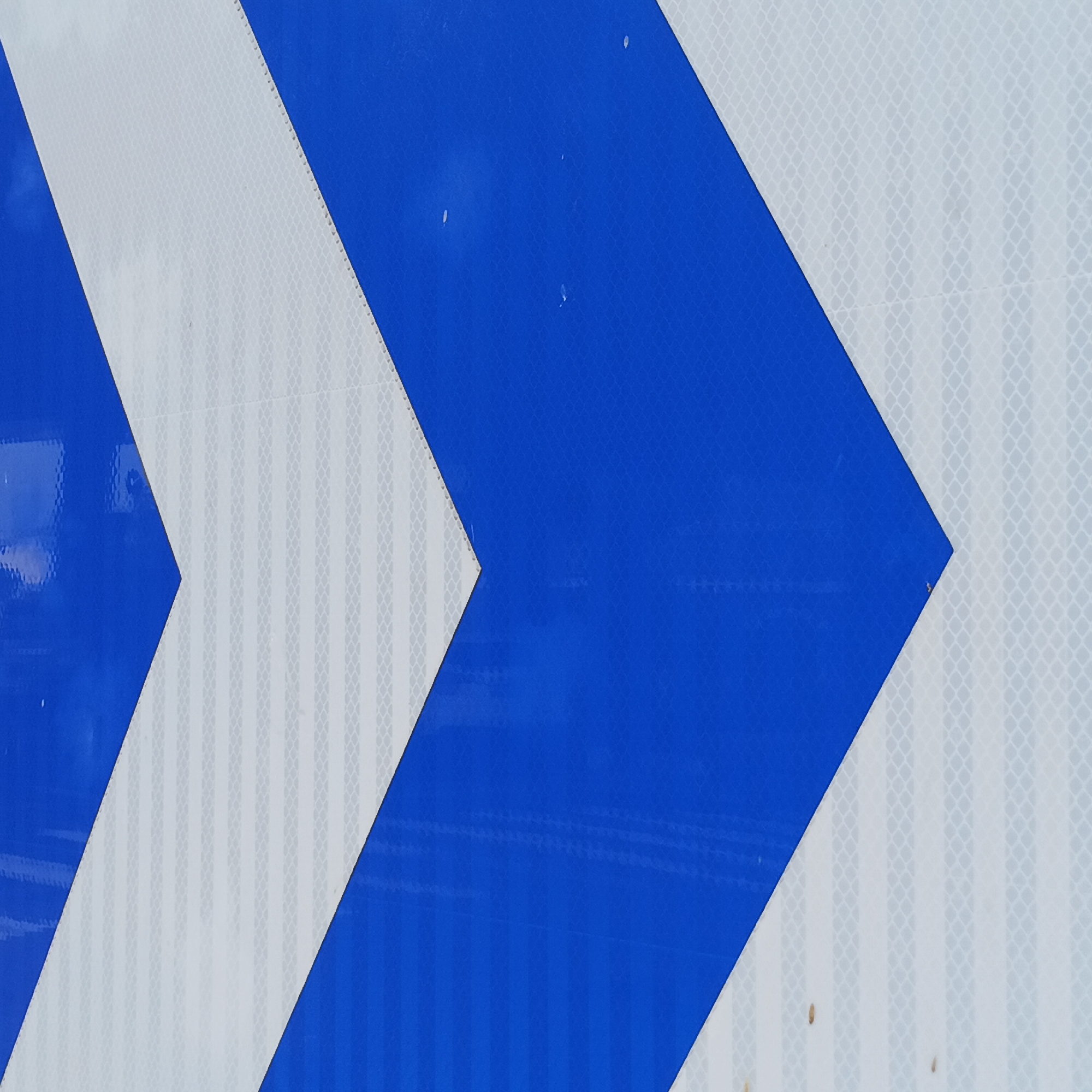 The image presents a close-up, abstract view of a reflective surface, likely part of a traffic sign or barrier. The primary subjects are alternating blue and white chevron-like patterns. The blue sections are solid and reflective, forming angular shapes that point towards the right side of the frame. The white sections are also highly reflective and display a fine, repeating grid or honeycomb texture, characteristic of retroreflective material.
No specific activity or event is depicted, as the image is a static shot of an object. Small imperfections, such as tiny white specks on the blue areas and brownish spots on the white sections, are visible, suggesting some wear or environmental exposure. There is no visible text. The time of day, weather, and specific location cues (beyond the provided Chancheng District, China context) are not discernible from the image itself. The composition highlights the geometric design and the material's property of reflecting light for visibility.
