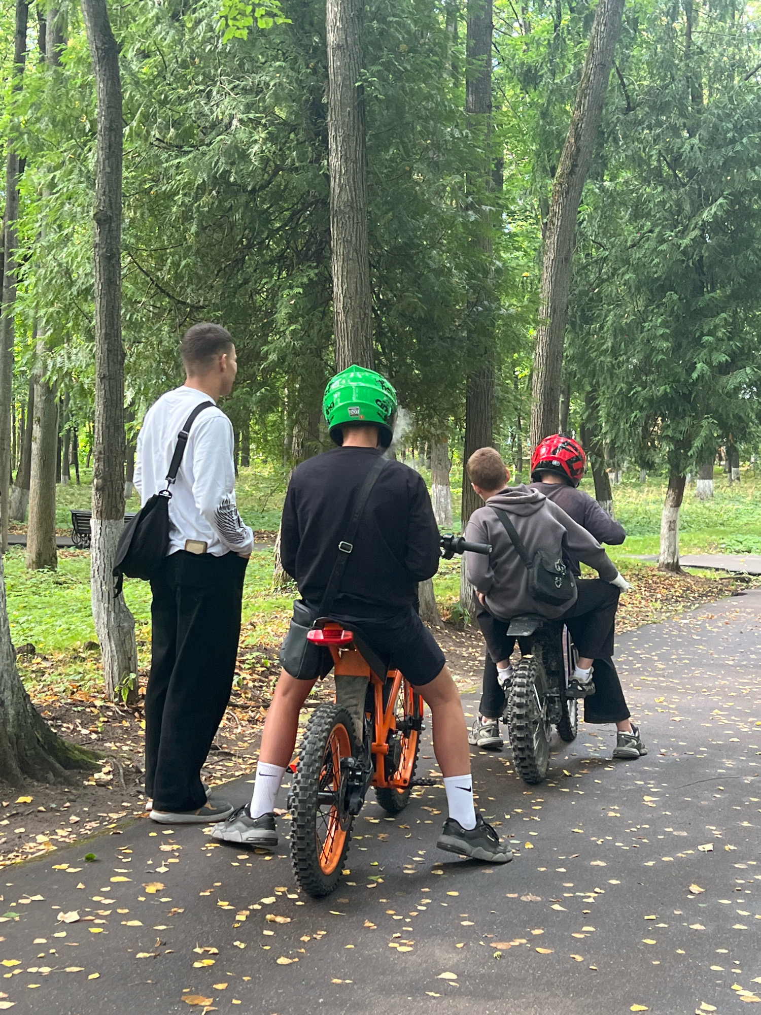 In Klin, Russia, four individuals are engaged in an outdoor activity in a park-like setting. Two people are seated on motorcycles, one with a red helmet and another with a gray hoodie. A third person, wearing a green helmet, is also on a motorcycle, and appears to be smoking. The fourth person, in a white long-sleeved shirt and black pants, stands beside them. The scene is characterized by tall trees with green foliage, and a paved path covered with scattered fallen leaves.