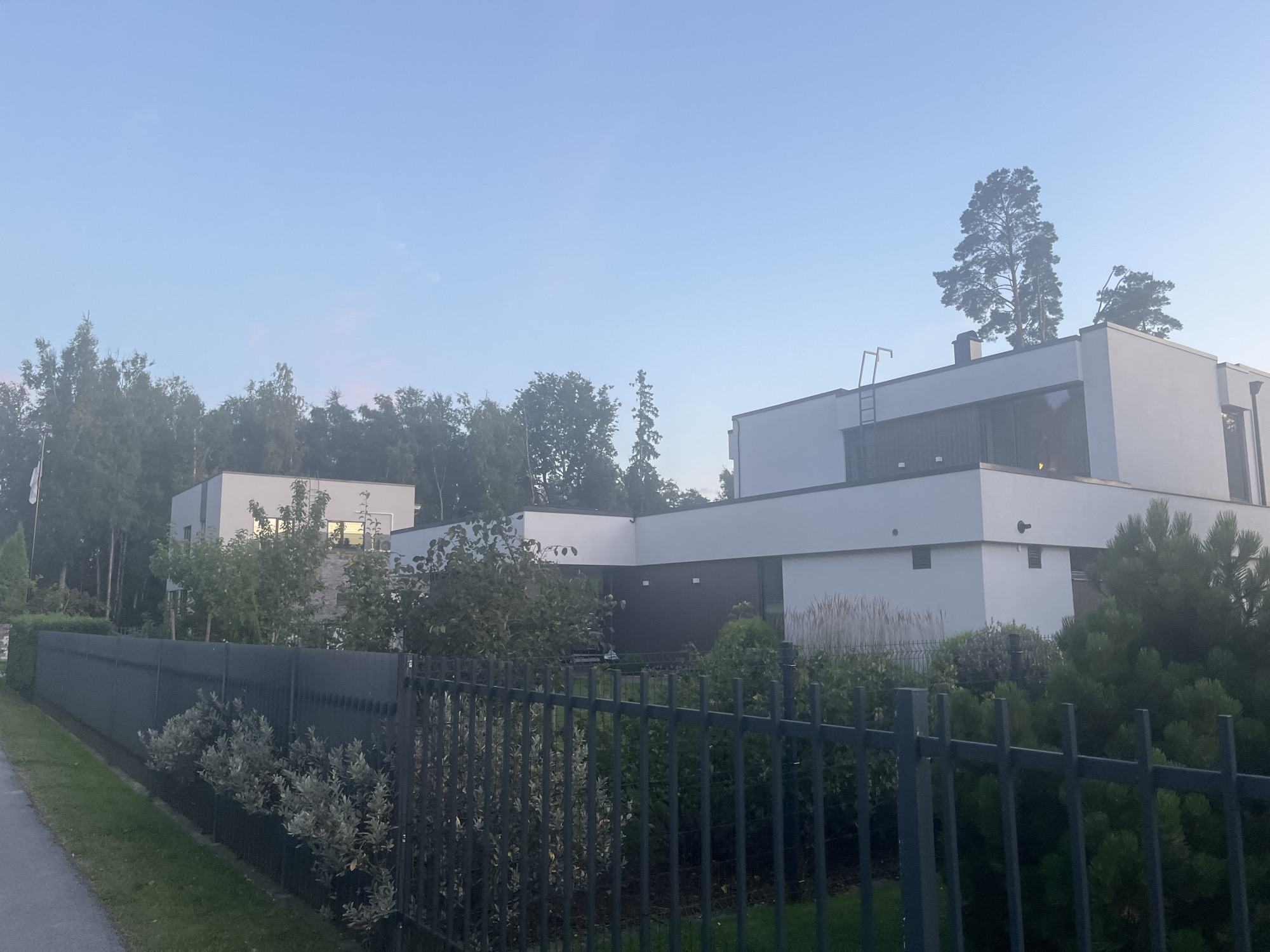 The image depicts a modern, two-story white house with large windows and a flat roof, situated in the city of Haabneeme alevik, Estonia. The house is surrounded by lush green trees and manicured bushes, with a dark metal fence separating the property from a paved walkway. The sky is a clear, pale blue with a hint of pink, suggesting it is either early morning or late evening. A flagpole is visible on the left side of the image, and a ladder is attached to the side of the house. The overall scene conveys a sense of tranquility and affluence.